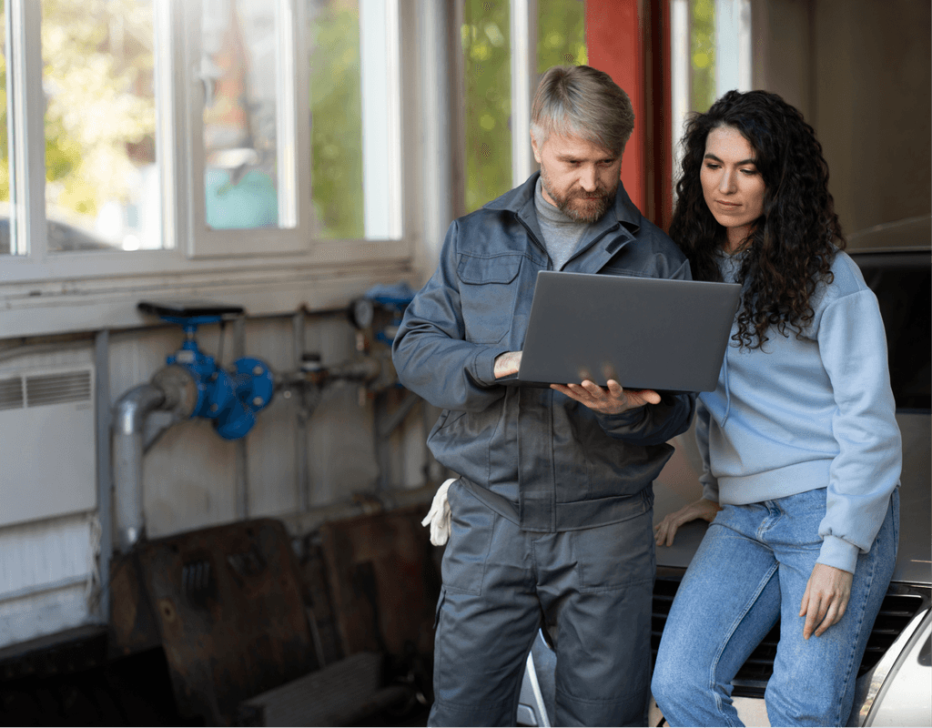 Auto mechanic showing a customer a laptop in a garage. They are looking at the screen, discussing car repair.