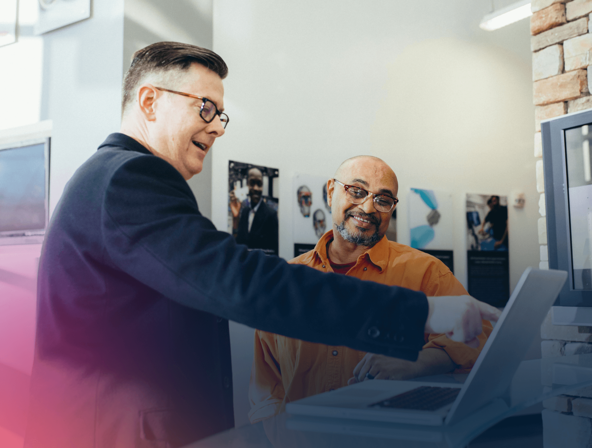 Man with glasses pointing at something on a laptop while talking to another man