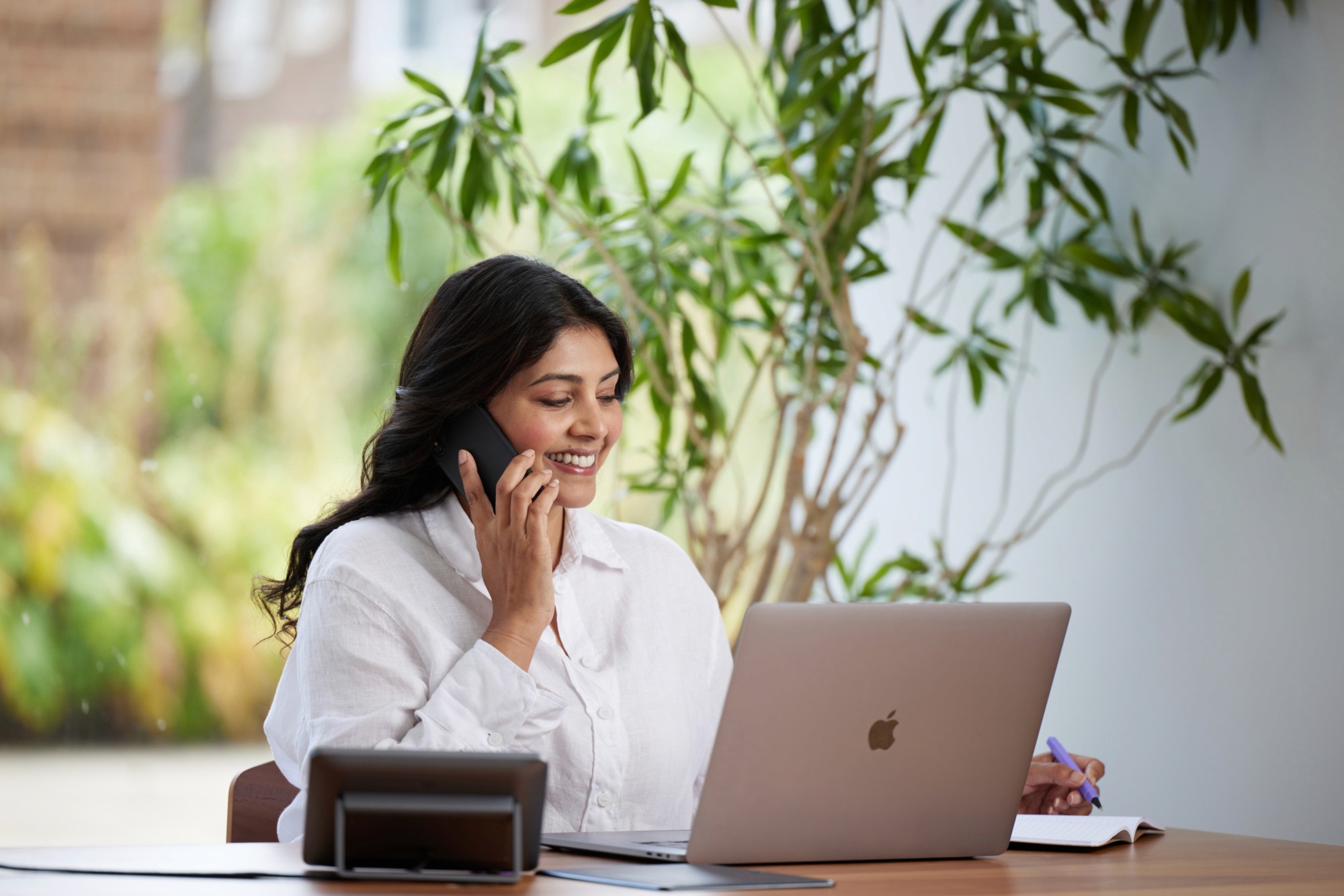 Woman smiling while talking on a phone and working on a laptop, taking notes at a desk in a bright, plant-filled workspace.