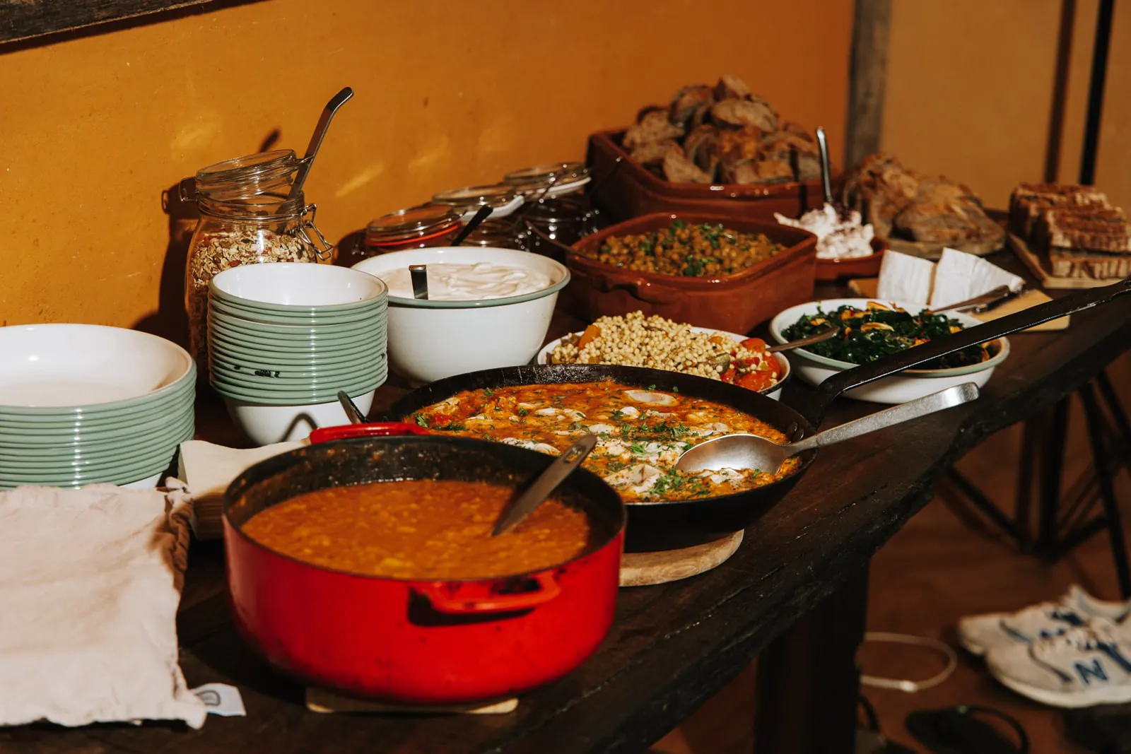 Een houten tafel vol eten: een rode pan soep, schalen met groenten en granen, brood en stapels borden