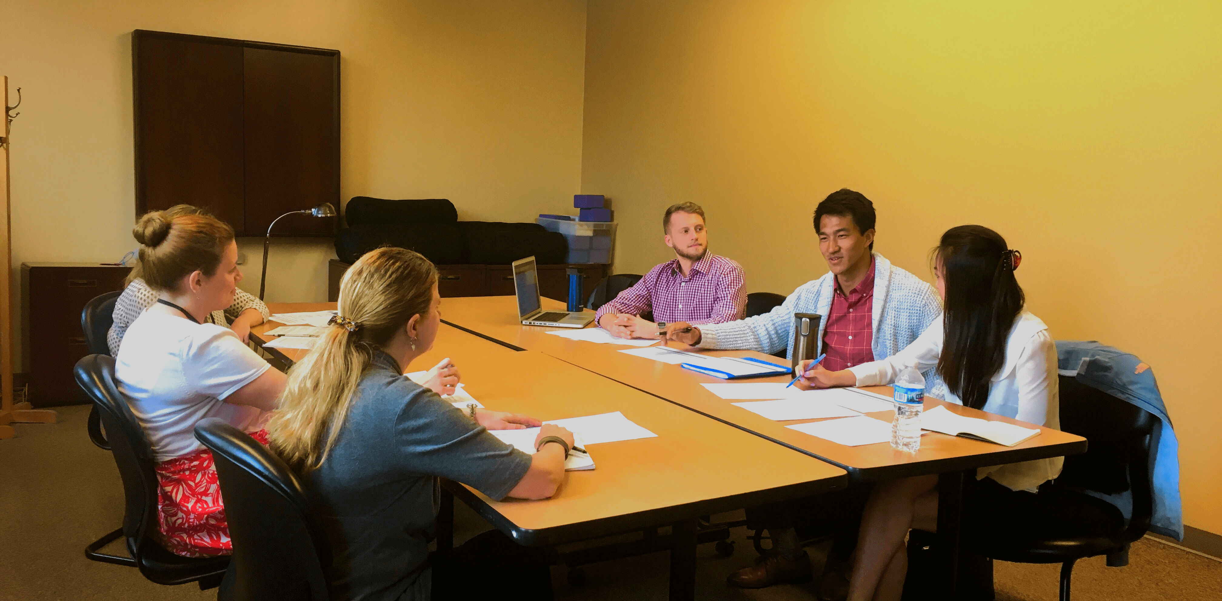 A group of six people sitting around a table in a meeting room engaged in discussion.