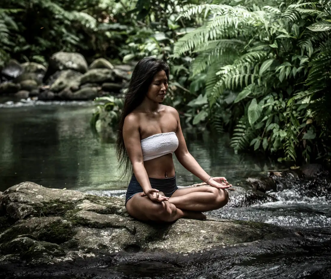 Woman meditating in nature during a yoga retreat in Bali, sitting on a rock in a river surrounded by jungle.