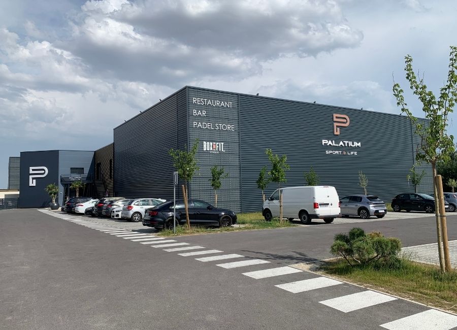 Bâtiment achevé avec façade texturée en métal ondulé, arboré à l'avant et allée bitumée.