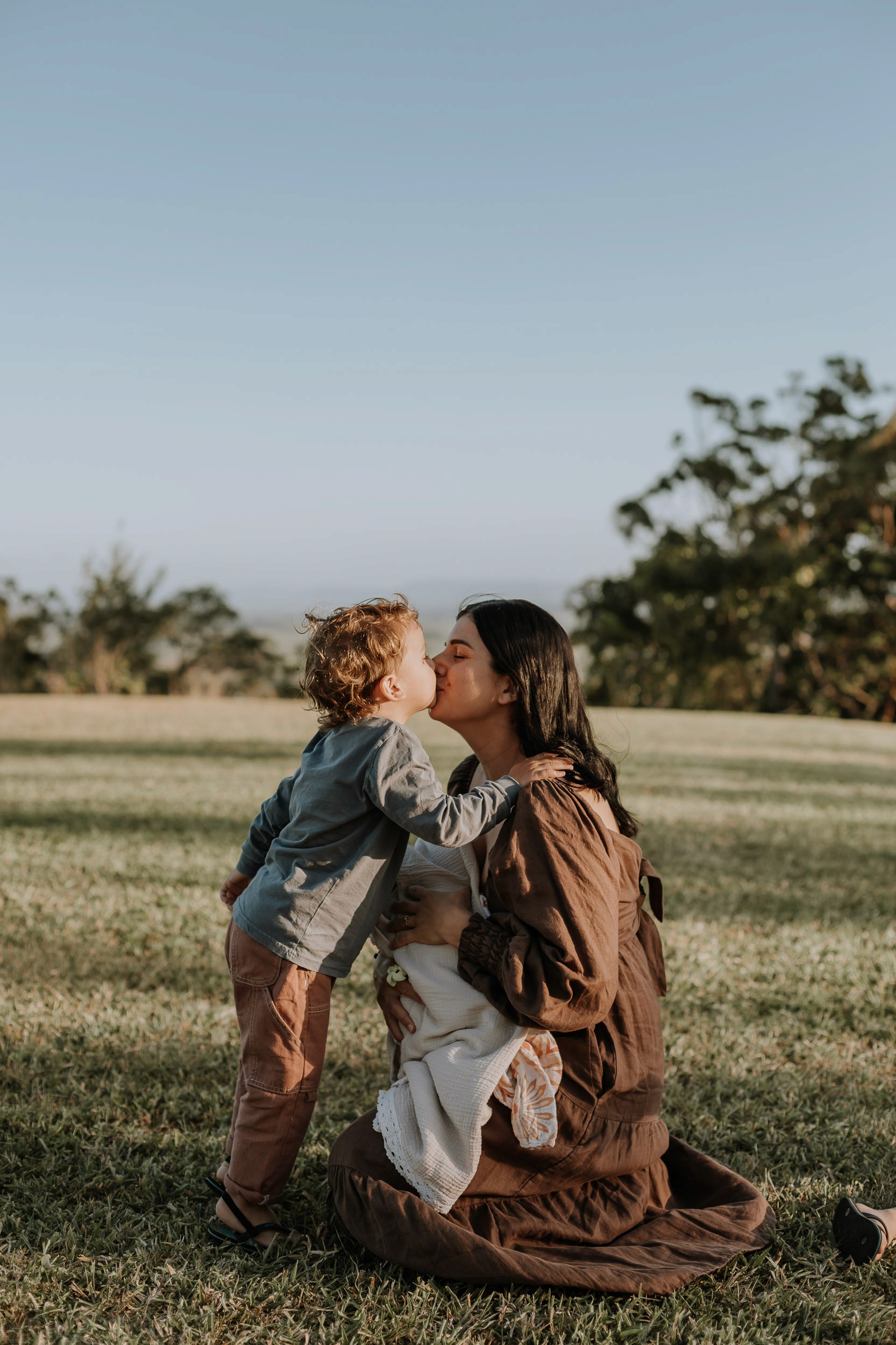 Relaxed candid family moment captured at golden hour with warm sunset light and child giving mum a kiss