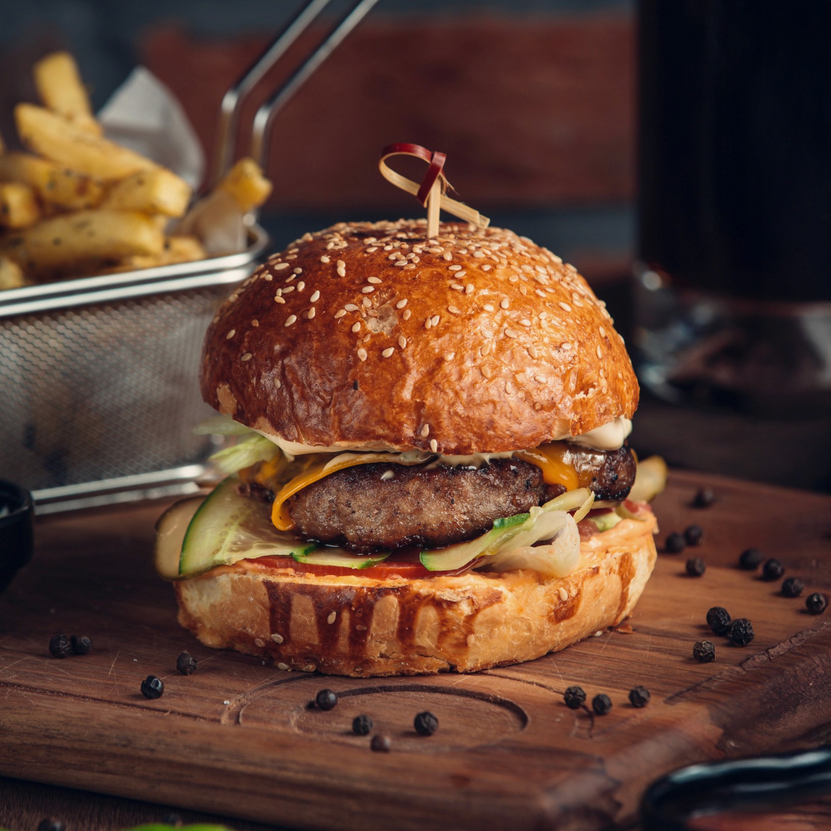 Close-up of a loaded hamburger on a wooden board with scattered peppercorns and a basket of fries in the background.