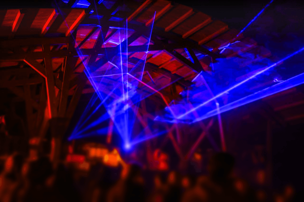 A person stands against a vibrant background of orange and yellow wavy patterns. They are wearing a dark coat, looking contemplative and thoughtful.