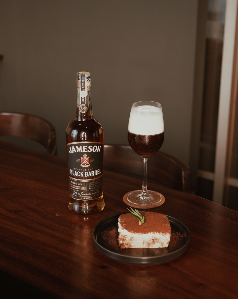 A bottle of Jameson Black Barrel whiskey next to a dessert on a wooden table, accompanied by a cocktail.
