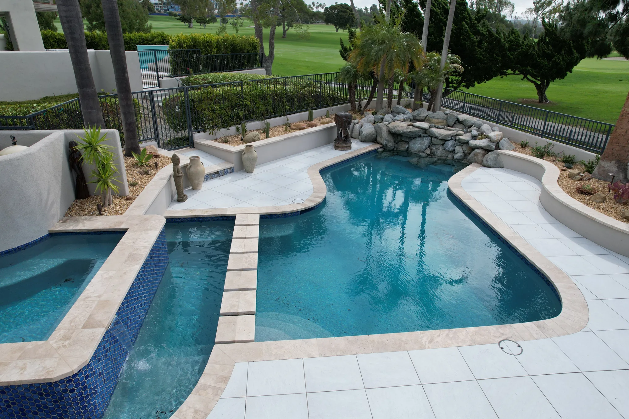 Curved pool surrounded by pale paving, with a raised blue-tiled spa and a rock water feature.