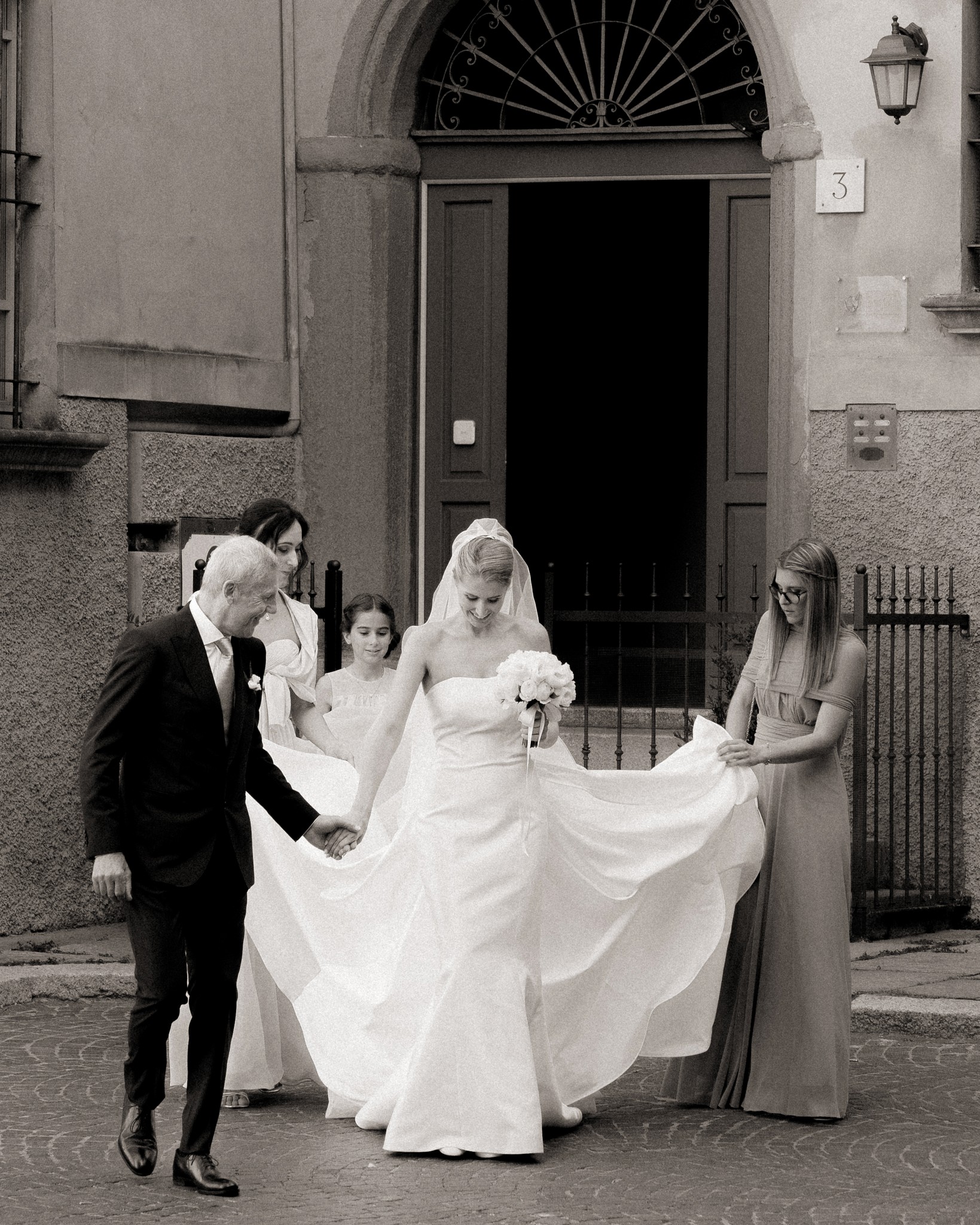 Sposa in abito bianco con velo, lo strascico sollevato da testimoni davanti al portone barocco di Villa Malenchini