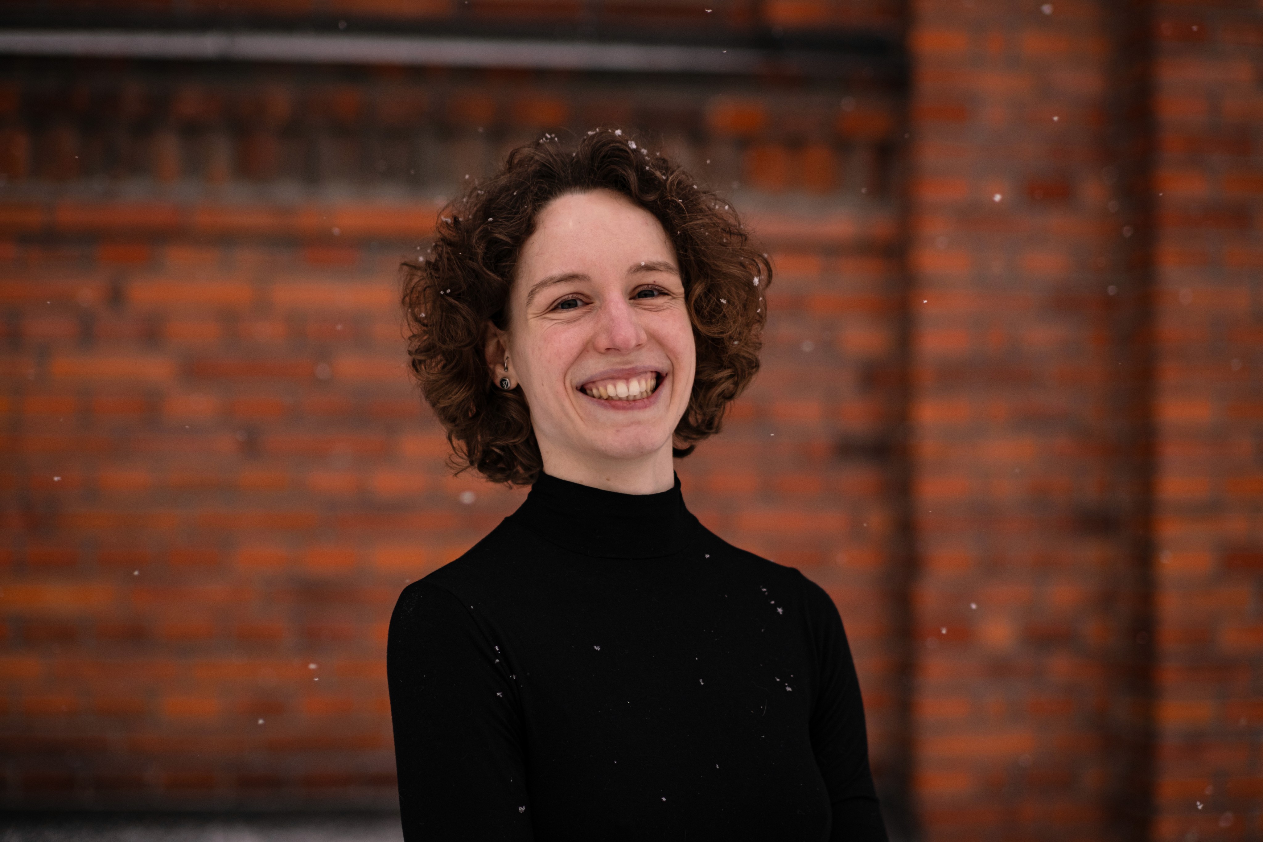 Medium close-up shot of a woman in her early thirties with curly hair and a black longsleeve, looking into the lens and smiling