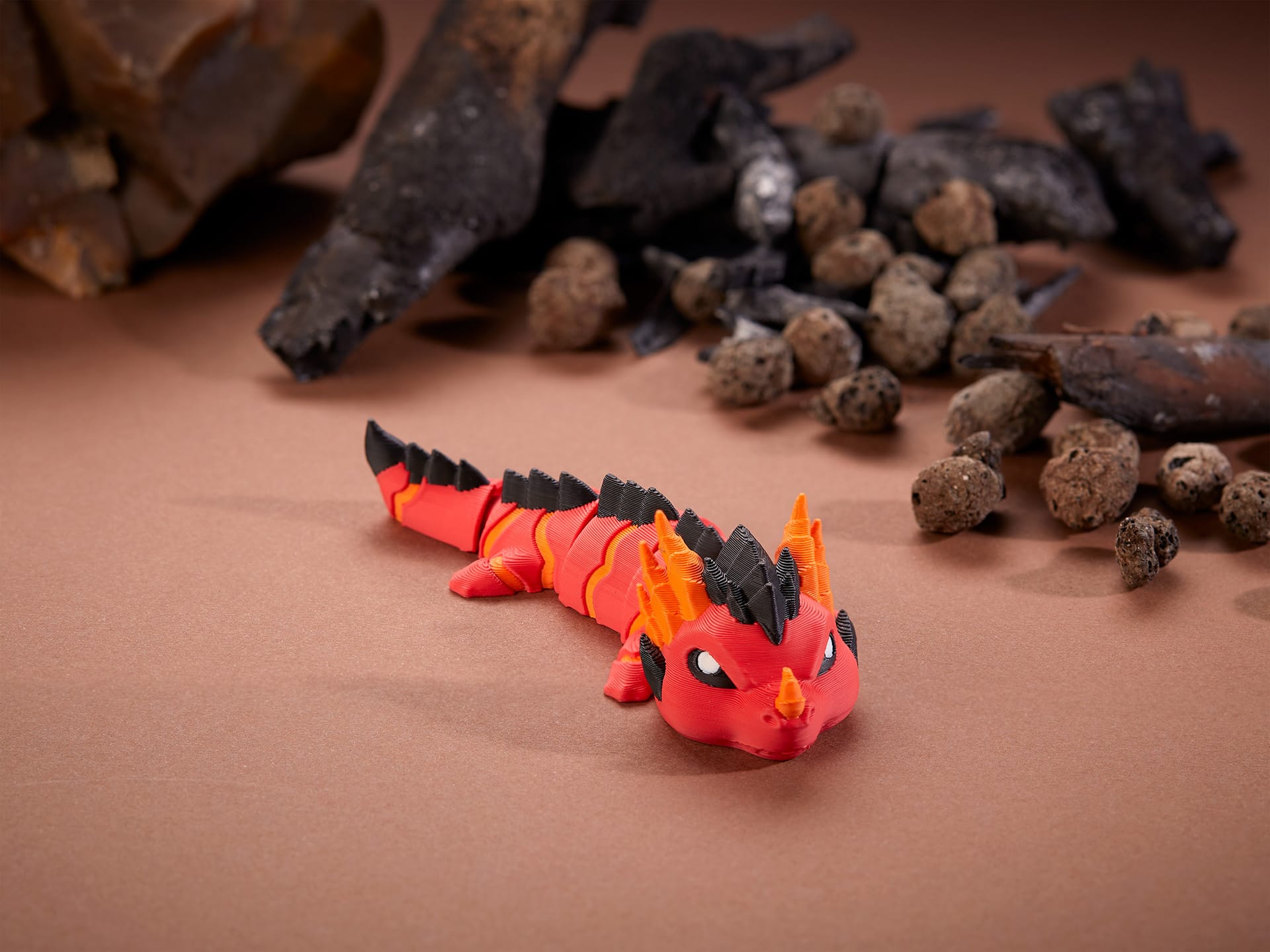 A small, intricately crafted red and black 3D print magma dragon with orange horns is set against a backdrop of scattered rocks and pieces of charred wood on a brown surface.