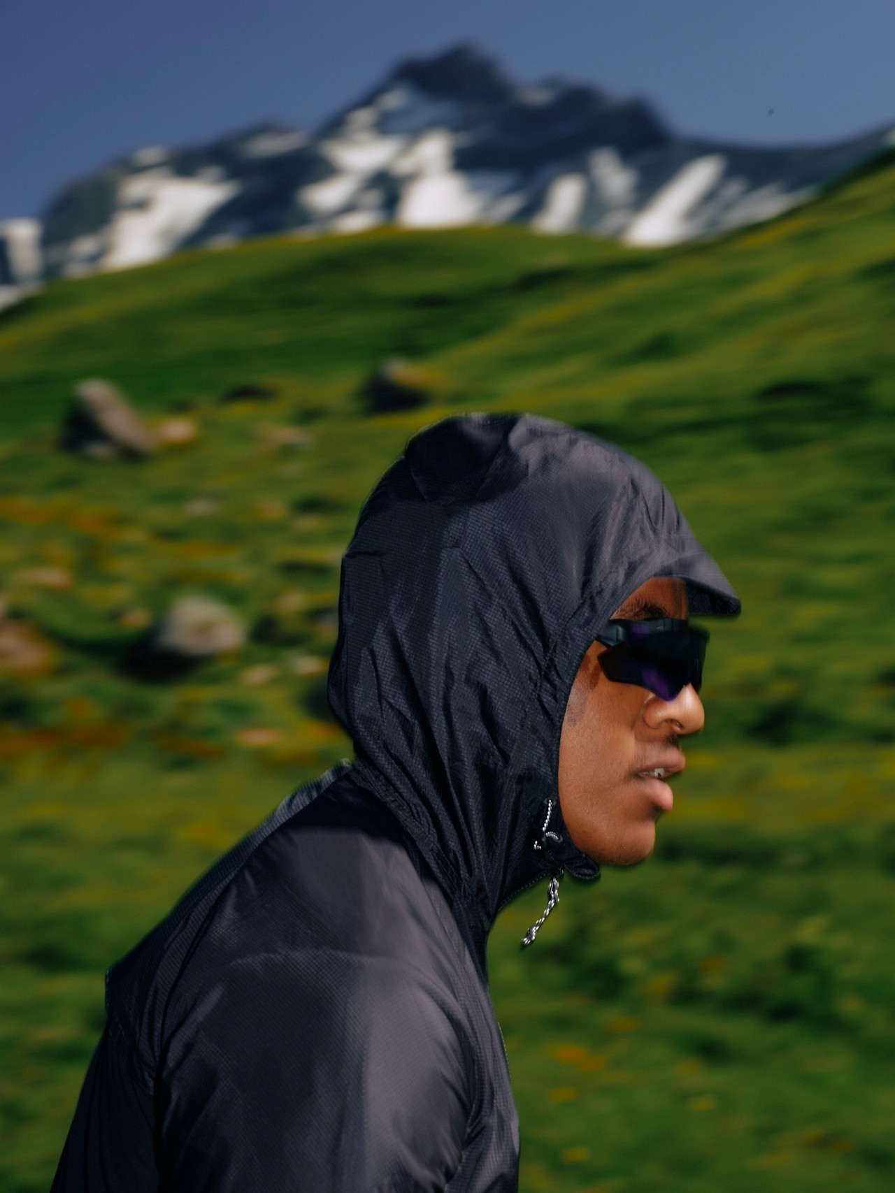 A person wearing a hooded jacket and sunglasses standing in a green mountainous landscape.