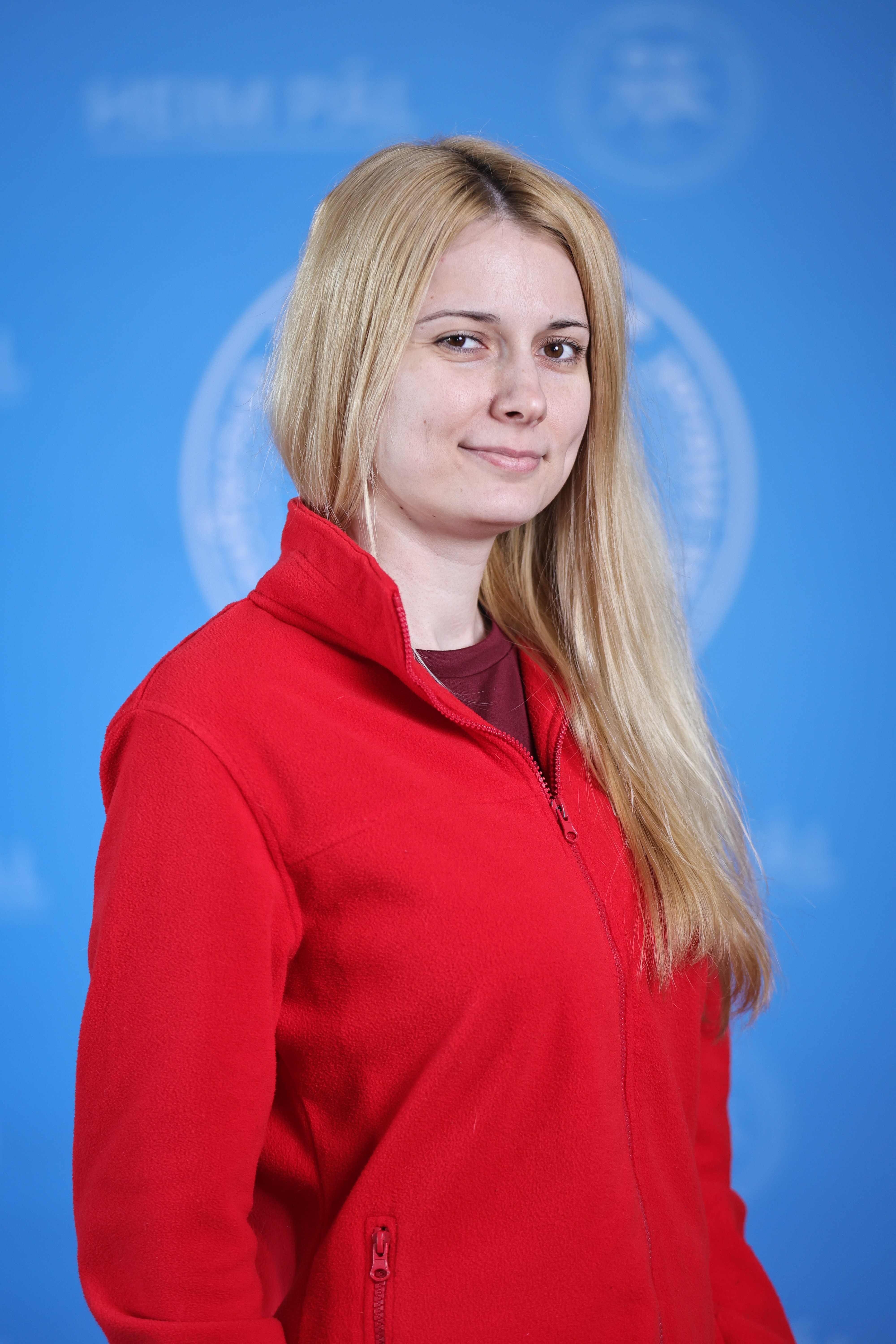 Hosszú, világos hajú nő mosolyogva áll egy hivatalos, kék háttér előtt, piros ruhában.