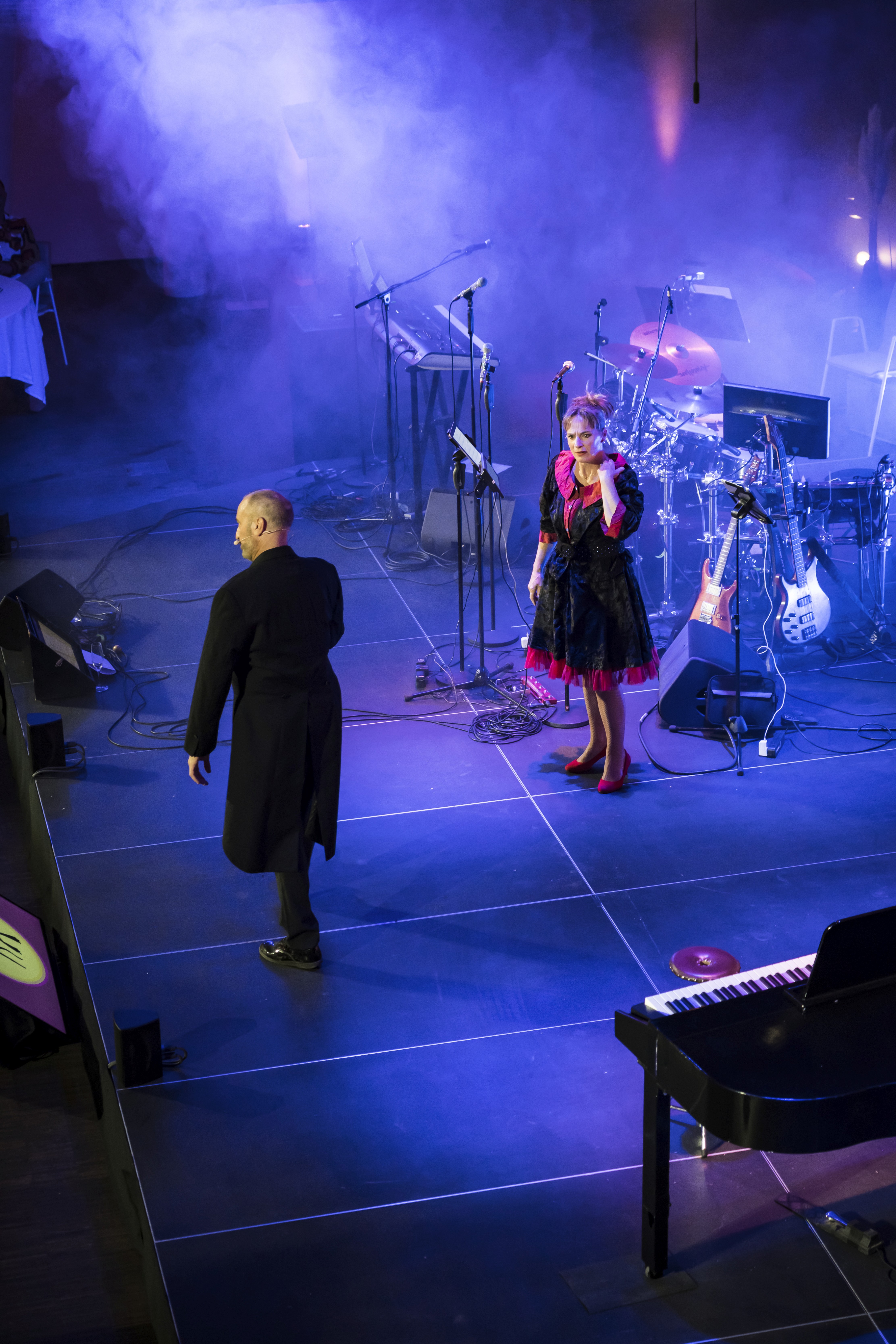 Das Foto zeigt eine dramatische Szene auf einer Bühne, die in tiefes blaues und violettes Licht getaucht ist und von starkem Bühnennebel durchzogen wird. Im Zentrum stehen zwei Personen: Auf der linken Seite sieht man einen Mann von hinten, der einen langen, schwarzen Mantel trägt und sich in Richtung der zweiten Person bewegt. Rechts steht eine Frau, die ihm zugewandt ist. Sie trägt ein dunkles, theatralisches Kleid mit auffälligen pinkfarbenen Akzenten am Kragen und am Saum sowie rote Schuhe. Im Hintergrund der Bühne ist das unbesetzte Equipment einer Live-Band aufgebaut, darunter ein Schlagzeug, Keyboards, E-Gitarren auf Ständern und diverse Mikrofone. Im rechten Vordergrund ragt zudem die Kante eines schwarzen Klaviers oder Flügels leicht ins Bild.
