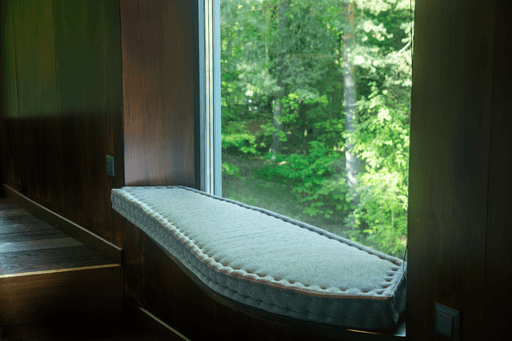 A minimalist interior with a window overlooking lush greenery, featuring a simple, padded bench.