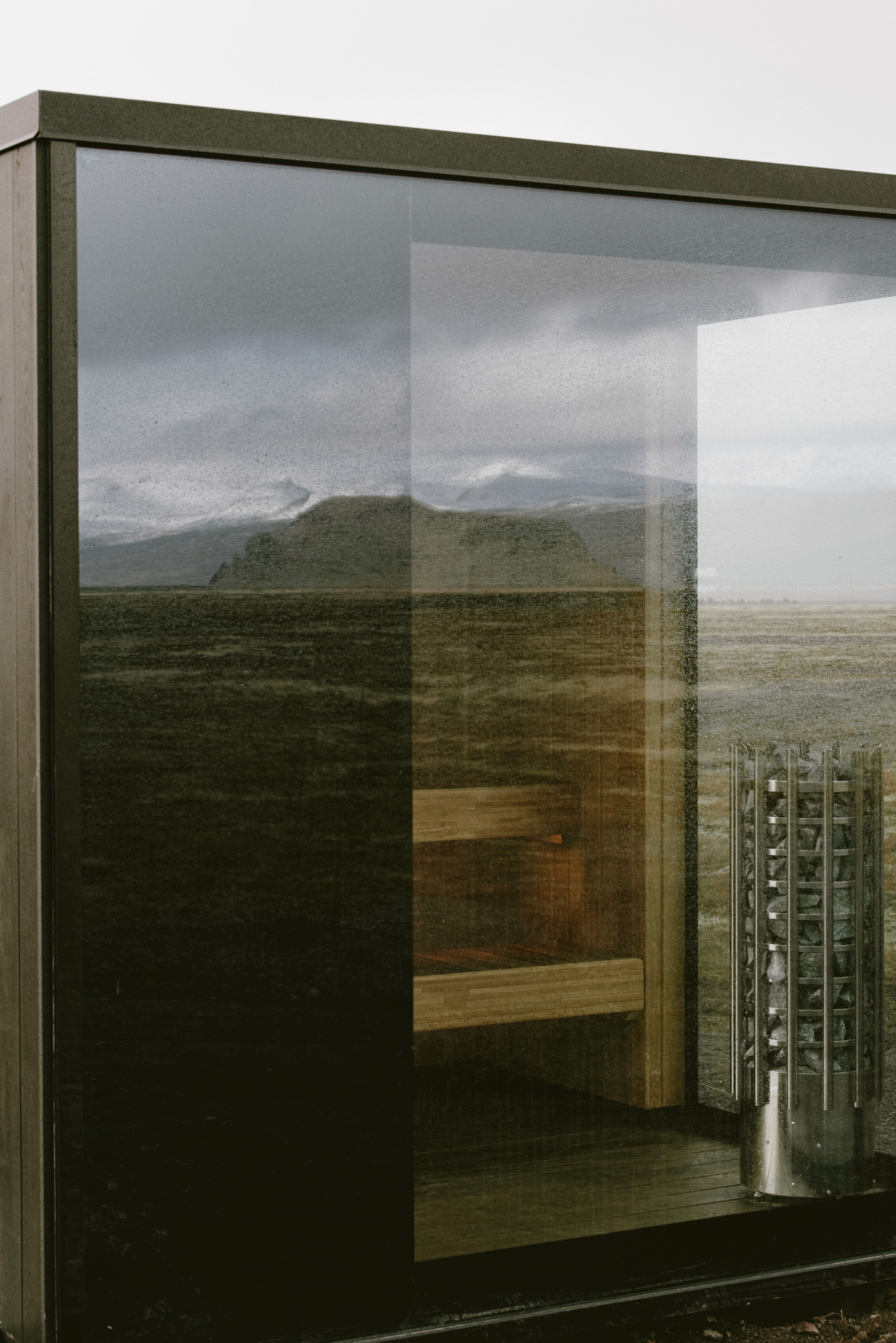 Glass-walled sauna in a modern luxury cabin, featuring timber interiors and panoramic views across a dramatic open landscape and distant mountains.
