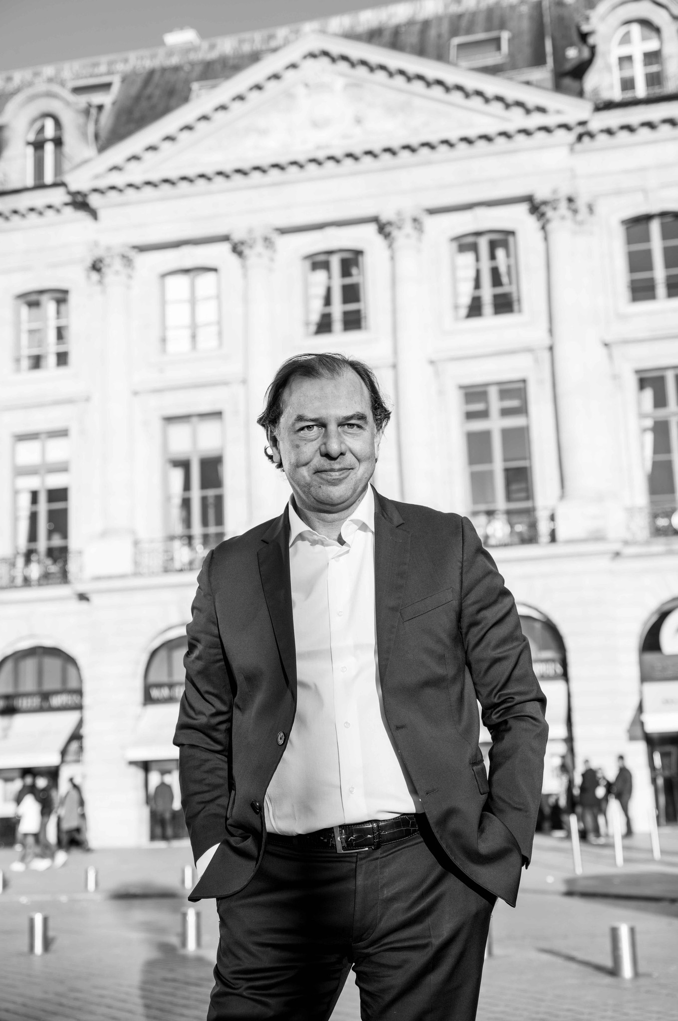 A smiling man with curly hair is sitting at a desk, looking at the camera, with a laptop and a lamp in the background.
