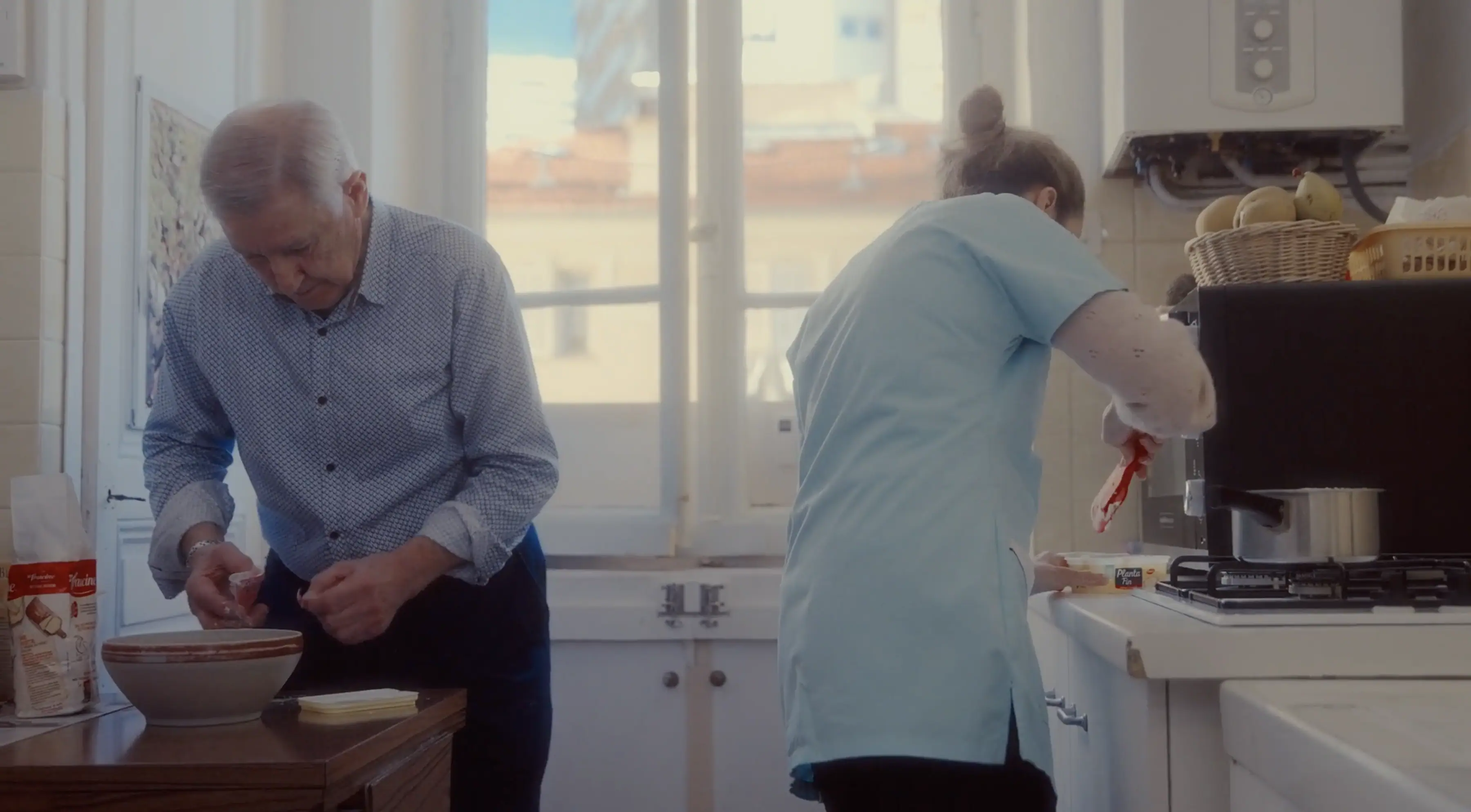 An elderly man and a caregiver are busy in a cozy kitchen, the man preparing ingredients at a table while the caregiver attends to a task by the stove, with sunlight streaming in through a large window, creating a warm and inviting atmosphere.