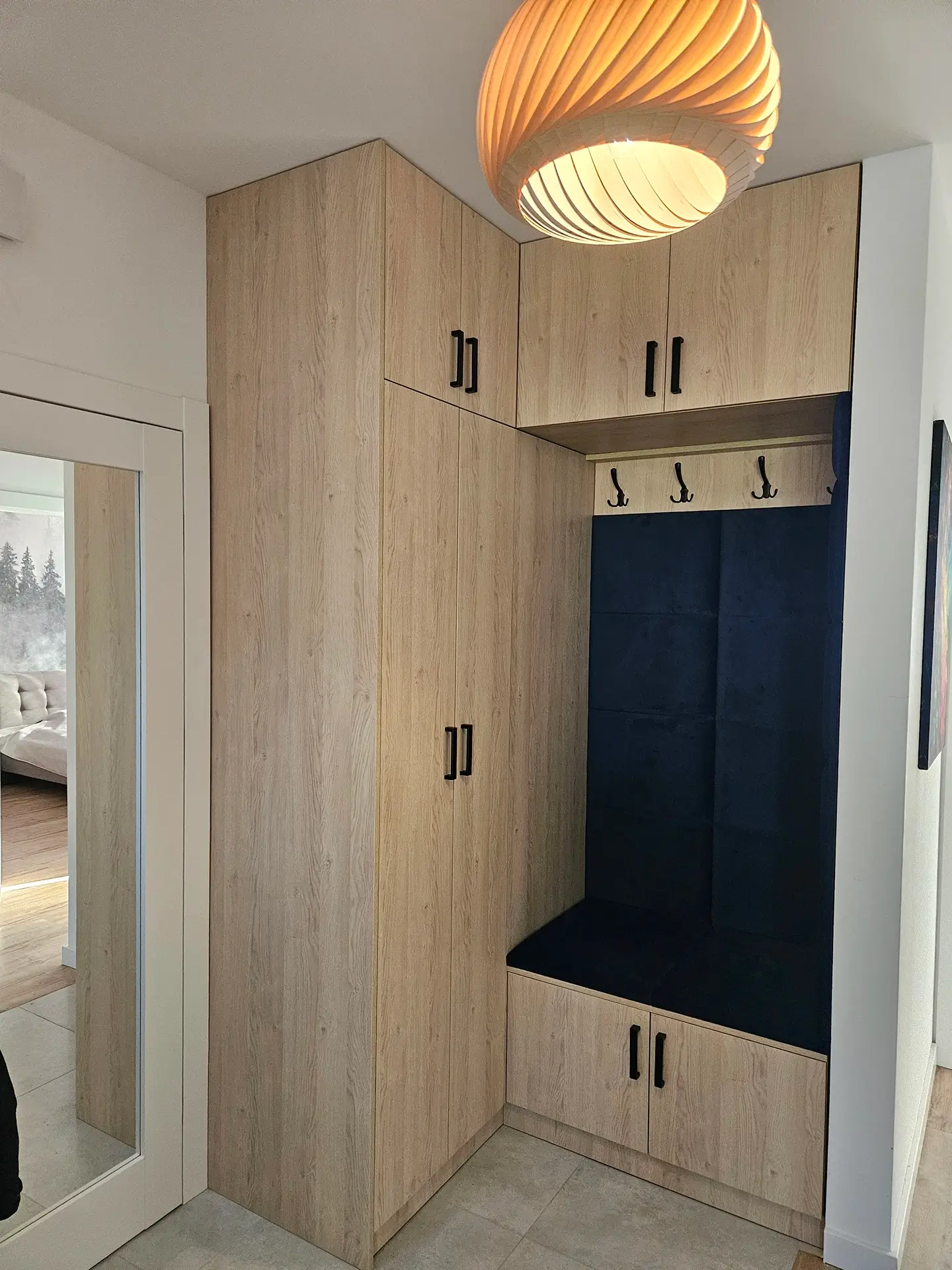 Zabudowa przedpokoju na wymiar w kolorze drewna: szafa, siedzisko z tapicerką i wiszącą lampą.