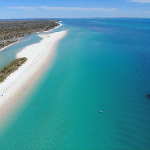 Aerial view of a pristine beach with turquoise waters, white sand, and a small kayak offshore. Sparse vegetation lines the shore.