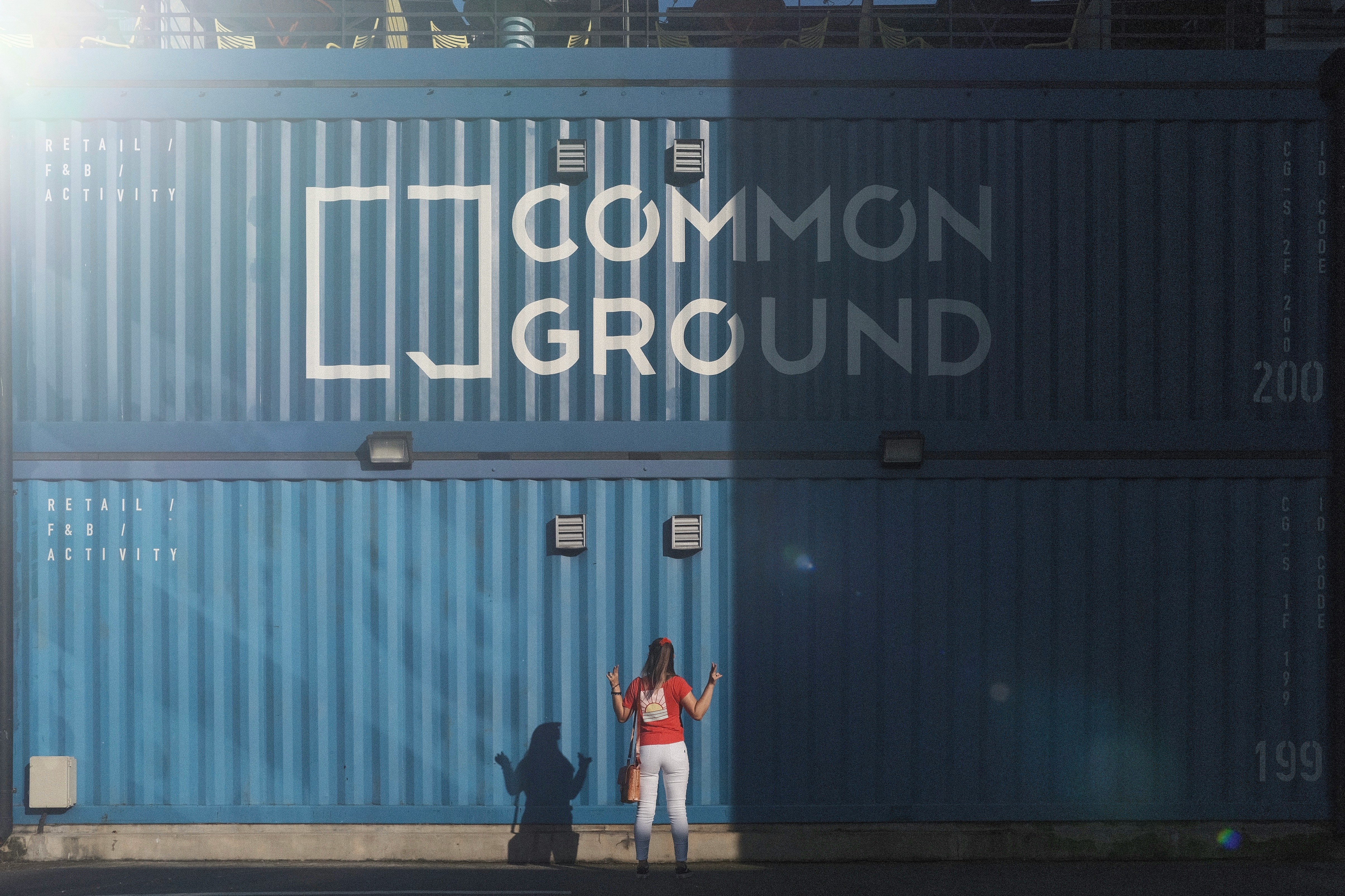 A blue shipping container with "COMMON GROUND" text and logo painted on it. Two people's shadows are visible against the container in what appears to be sunlight