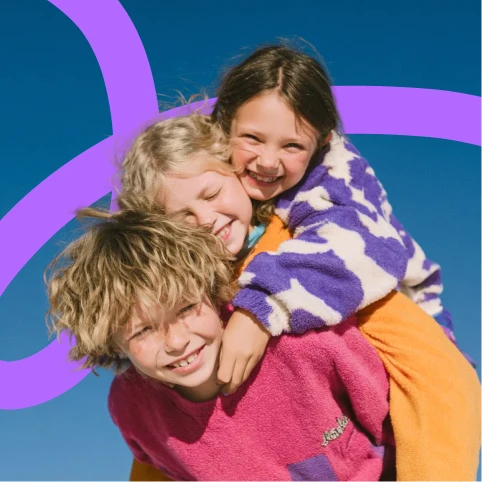 Three children smiling and playing together, one giving a piggyback ride, against a clear blue sky with a purple graphic line in the background.
