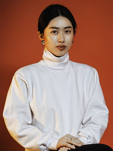 A person with dark hair pulled back is captured in a head-and-shoulders portrait against a solid orange-brown backdrop. They are wearing a white turtleneck long-sleeved shirt and have their arms folded across their chest. The individual's gaze is directed straight at the camera, and their expression is neutral. They wear small, dangling earrings and a ring is visible on their left ring finger. The lighting is even and illuminates the subject clearly.