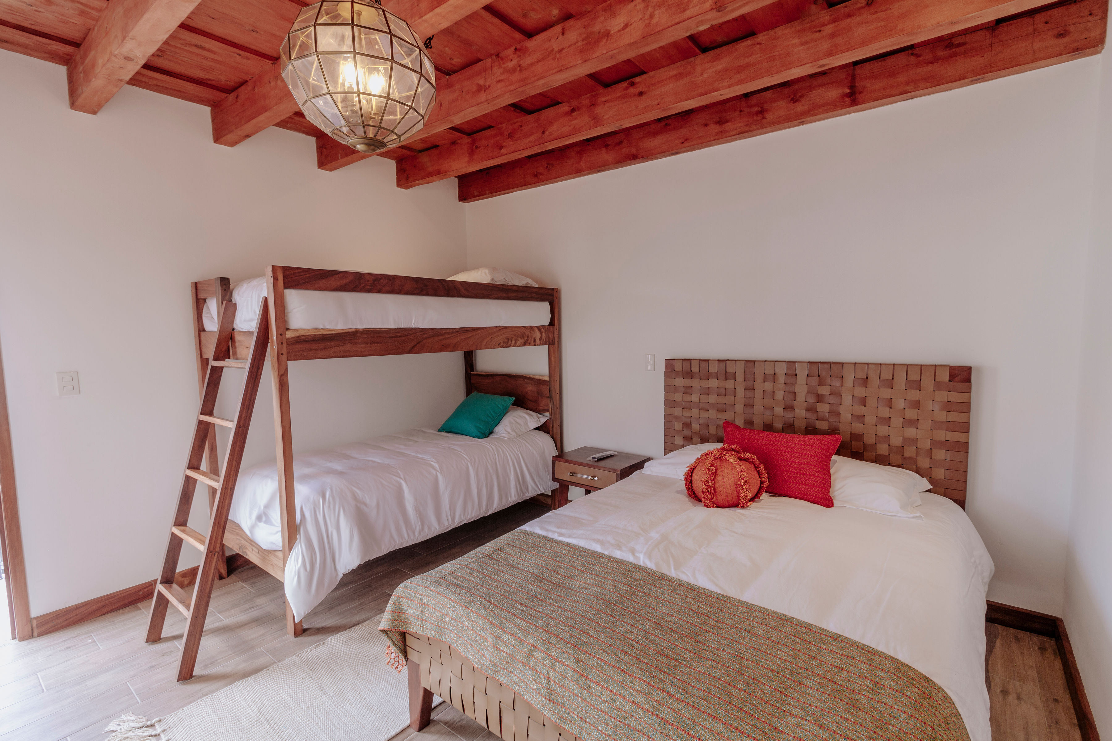 A cozy bedroom with a rustic wooden ceiling features a bunk bed with white linens and a single bed with earthy-colored bedding accented by red and orange pillows, adding warmth to the minimalist decor.