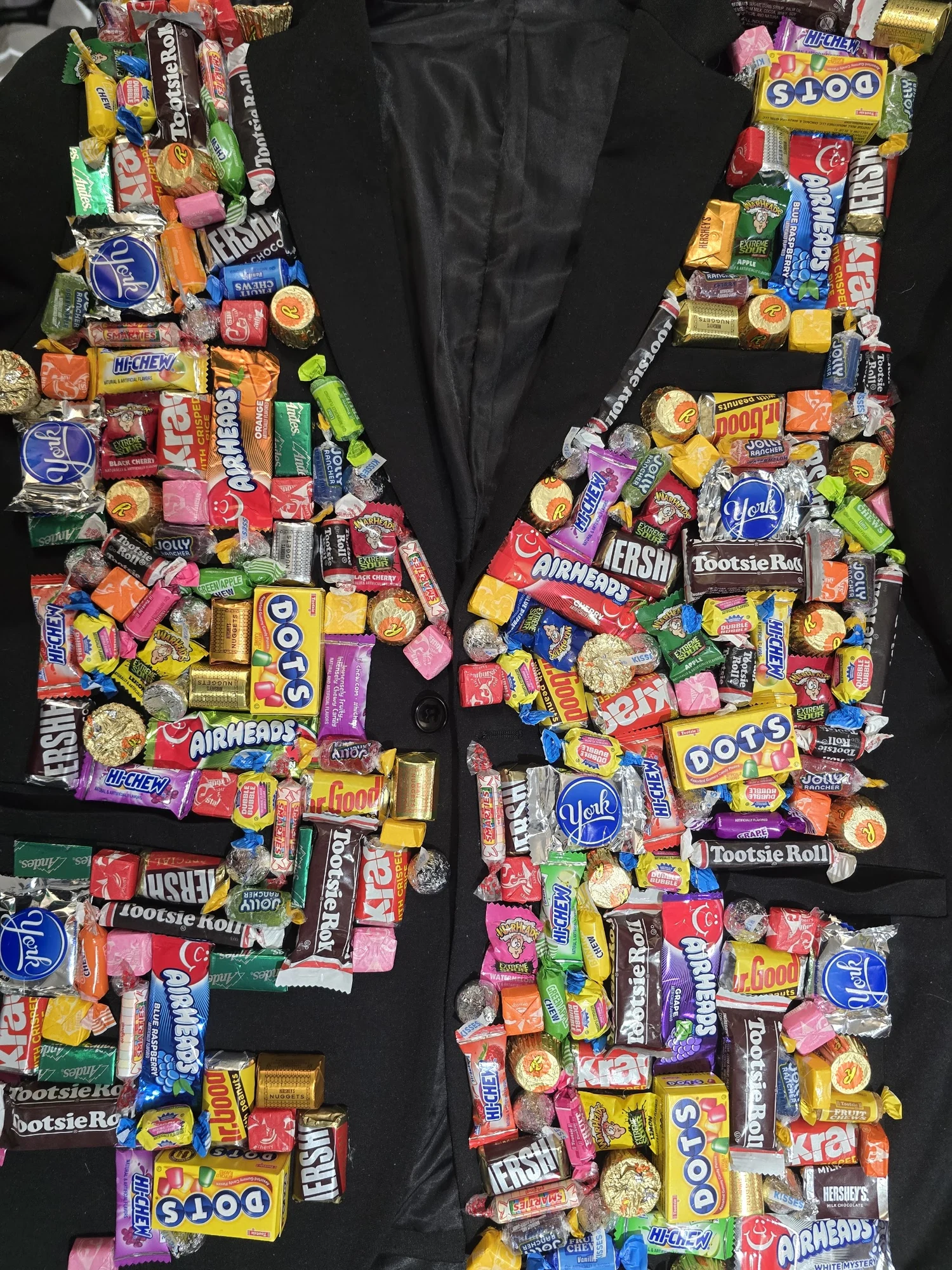 Direct-flash three-quarter portrait of No Amayume's black suit, densely covered in wrapped candy from lapel to sleeve.