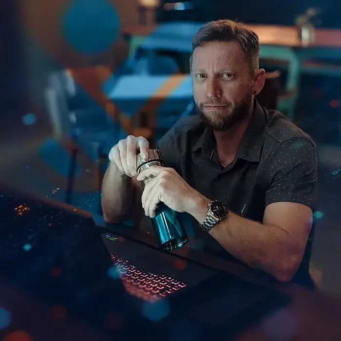 Imagem do Designer e Proprietário da Creativa UX Solutions, Bruno Nascimento, ele está sentado em sua mesa de trabalho em frente ao notebook com os cotovelos apoiados na mesa e abrindo uma garrafa metálica de água. Ele está vestido de camisa usa um relógio o ambiente tem tons de azul e laranja. 