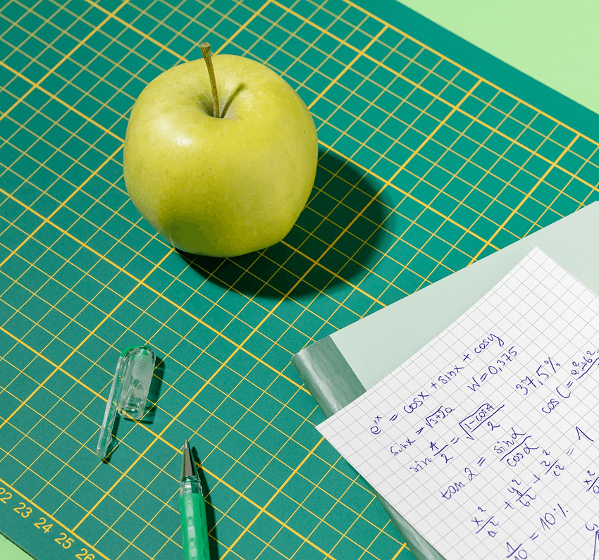 A green apple sits on a cutting mat with a pen and a sheet of handwritten notes beside it.