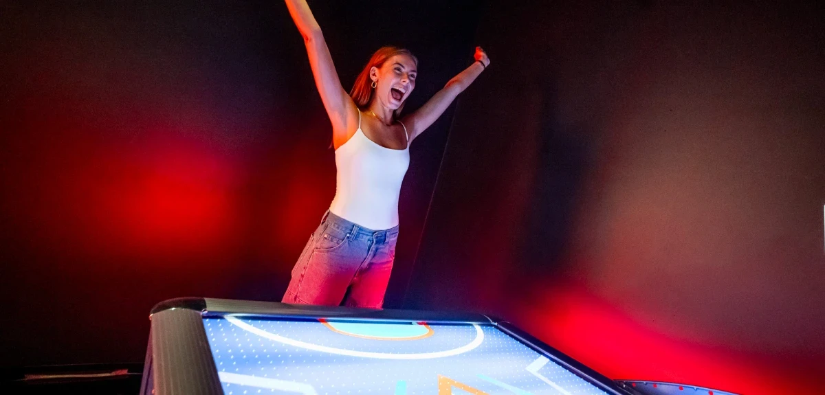 A woman celebrating with her arms up at a red lit air hockey table