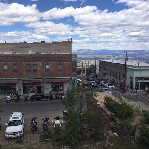 Улица в малък град с паркирани мотоциклети, автомобили и хора, които се разхождат. Сградите са от тухли, а на заден план се вижда живописен пейзаж.