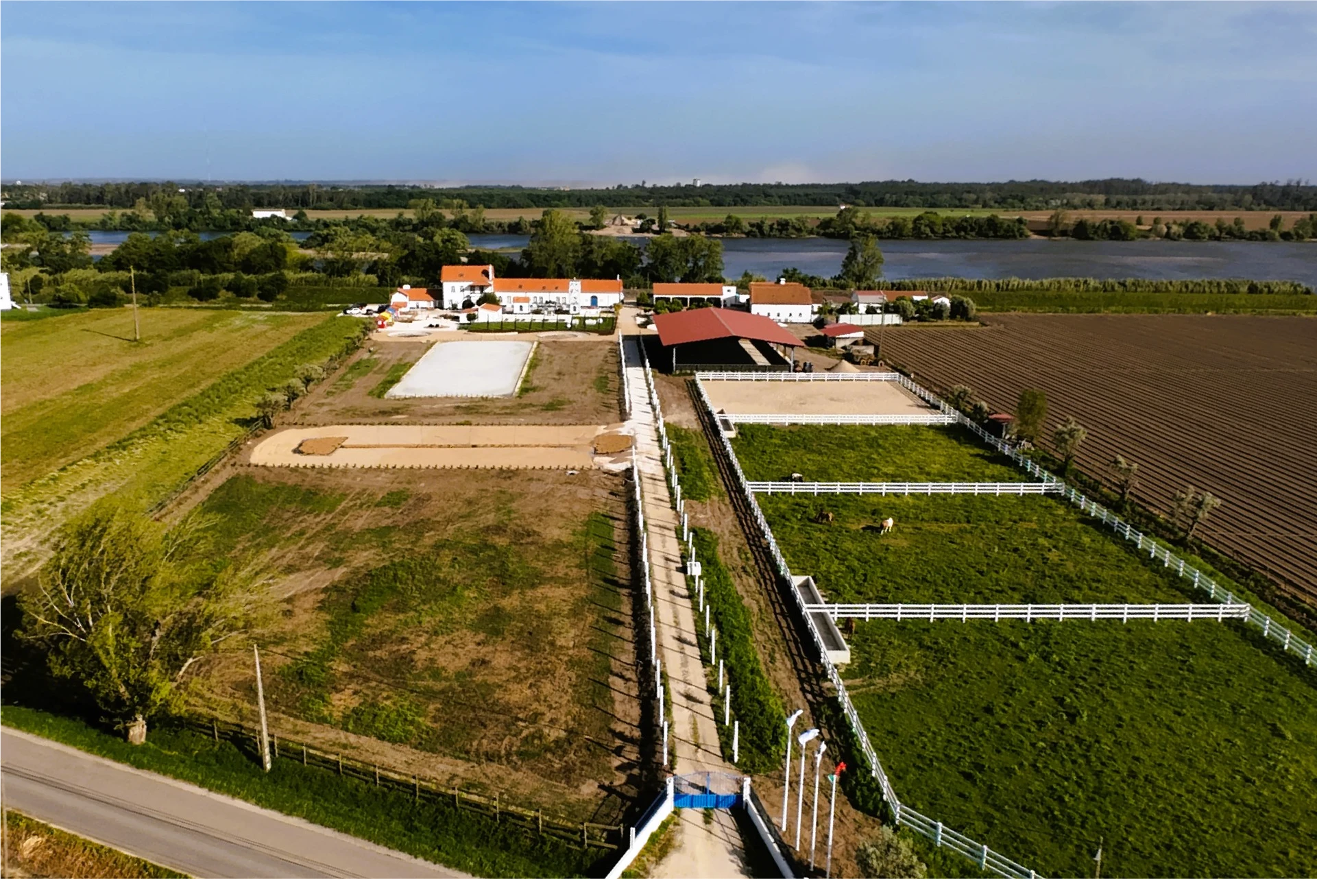 Aerial view of a spacious ranch with green fields, horse paddocks, and a central barn. A river and trees are visible in the background under a clear sky.
