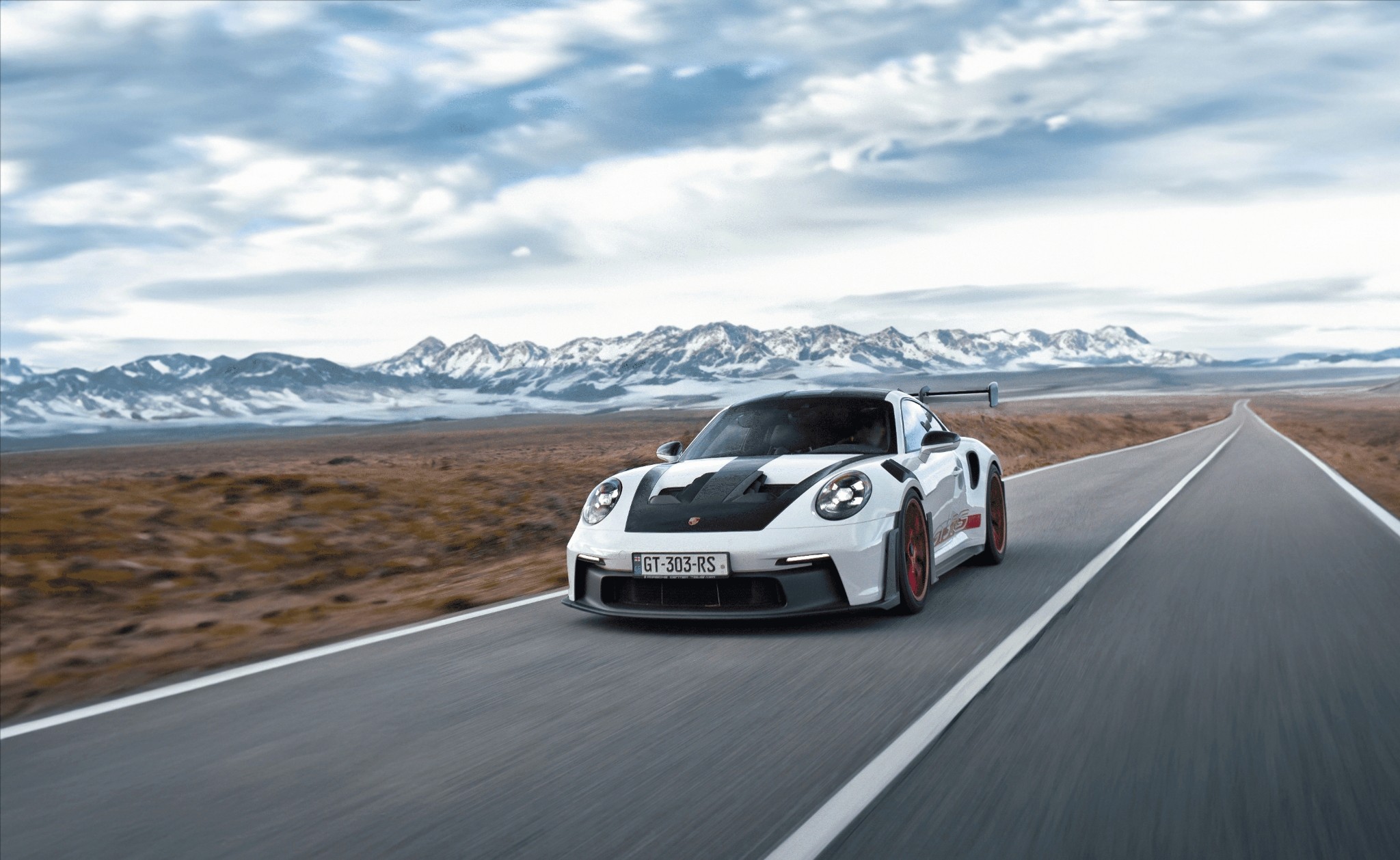 A sleek sports car with a white body and black accents speeds down an open highway, set against a backdrop of vast, rocky desert and distant snow-capped mountains under a cloudy sky.