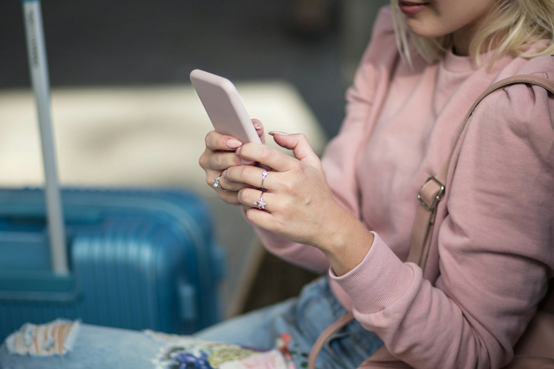 A woman seated with luggage beside her, holding a smartphone in both hands while wearing a pink sweater and jeans.
