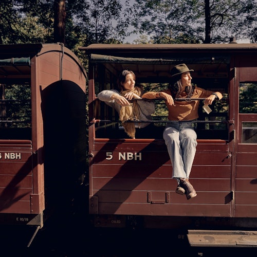 ਦੋ ਲੋਕ ਇੱਕ ਰੱਸੀ ਟ੍ਰੇਨ ਕਾਰ ਦੇ ਫੇਰੋਂ "5 NBH" ਲਿਖੇ ਬਾਹਰ ਝੁਕਦੇ ਹਨ, ਜਿਸਨੂੰ ਪਿੱਛੇ ਦਰੱਖਤਾਂ ਨੇ ਘੇਰਾ ਹੋਇਆ ਹੈ।