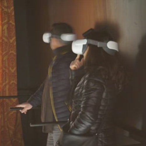 Due persone che indossano visori per la realtà virtuale in una stanza poco illuminata, in piedi vicino a una parete con una tenda decorata visibile.