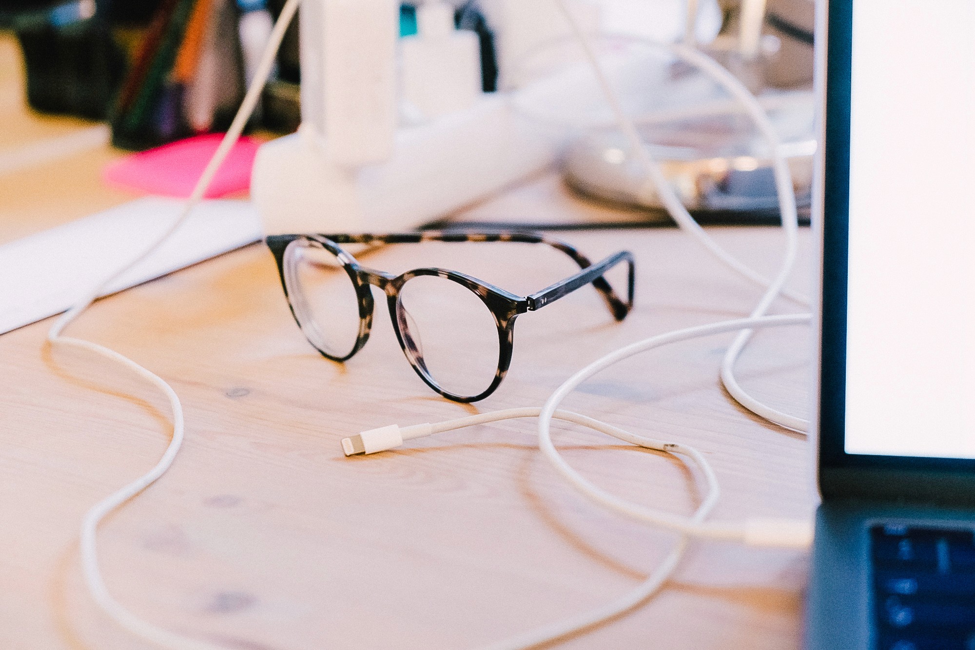 Brille mit dunklem Rahmen liegt auf einem Holztisch neben einem Ladekabel in einem modernen Büro.