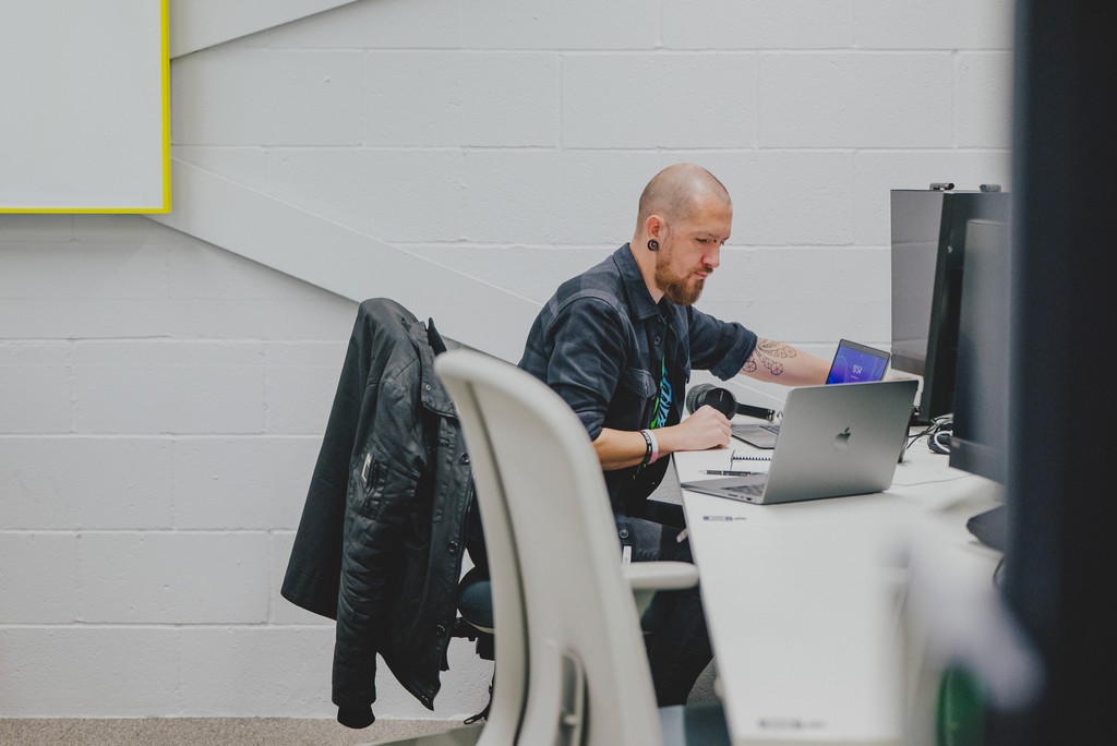 Person working at a desk in a modern office workspace, using a laptop and external monitors, with office chairs and a light‑coloured wall in the background.