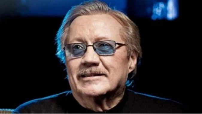 A man with light brown hair and a moustache is wearing blue-tinted glasses, captured against a dark background with subtle blue lighting for a sophisticated atmosphere.