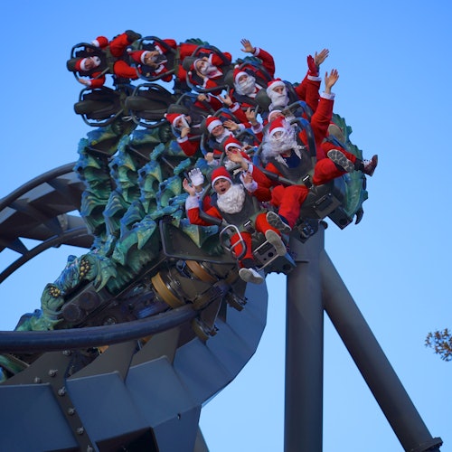 Eine Gruppe von Menschen in Weihnachtsmannkostümen fährt mit erhobenen Händen auf einer Achterbahn, vor einem klaren blauen Himmel.