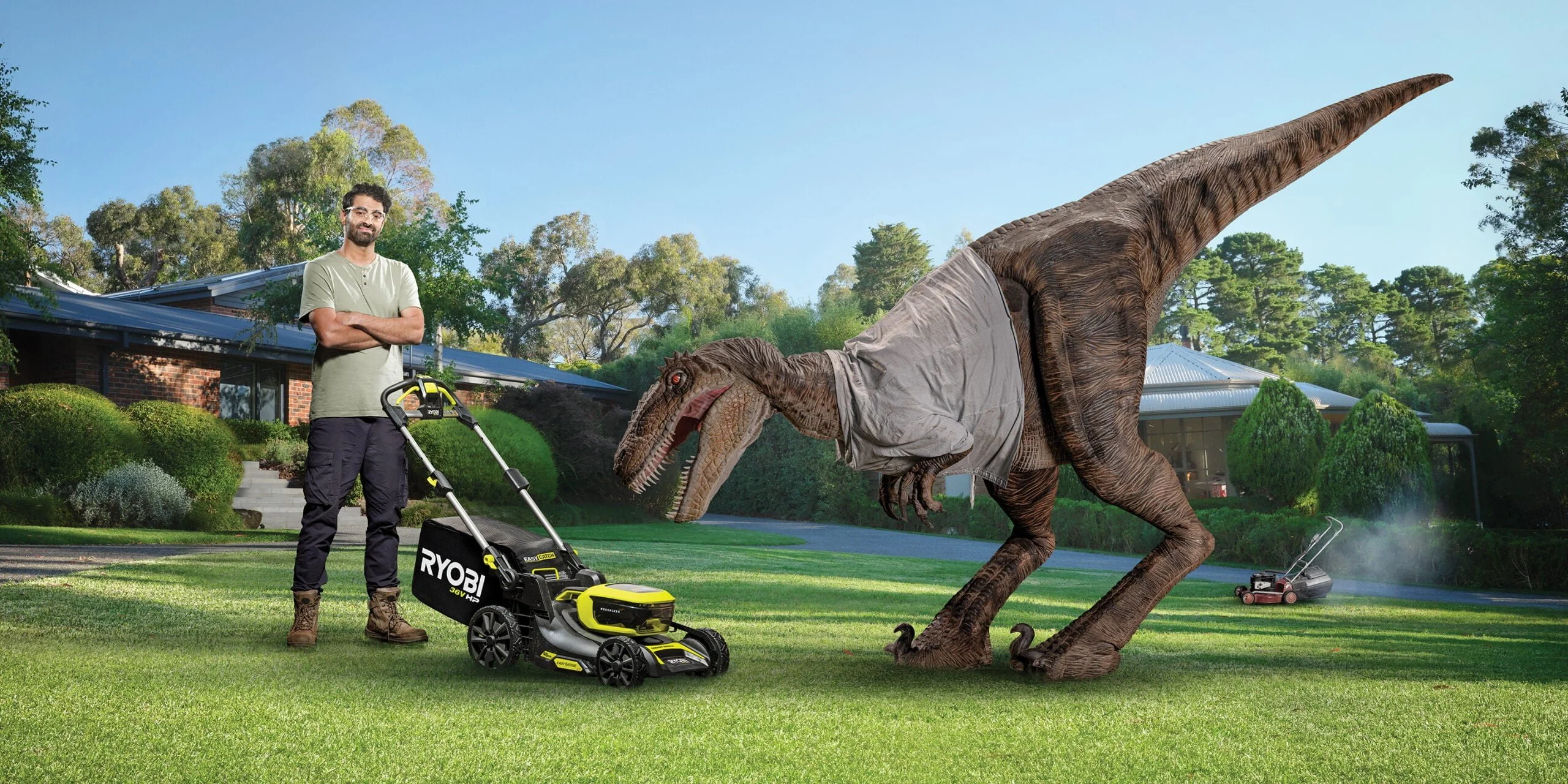 Man in safety glasses with a Ryobi battery mower facing a dinosaur wearing a shirt on a suburban lawn, from 'Petrol Evolved'