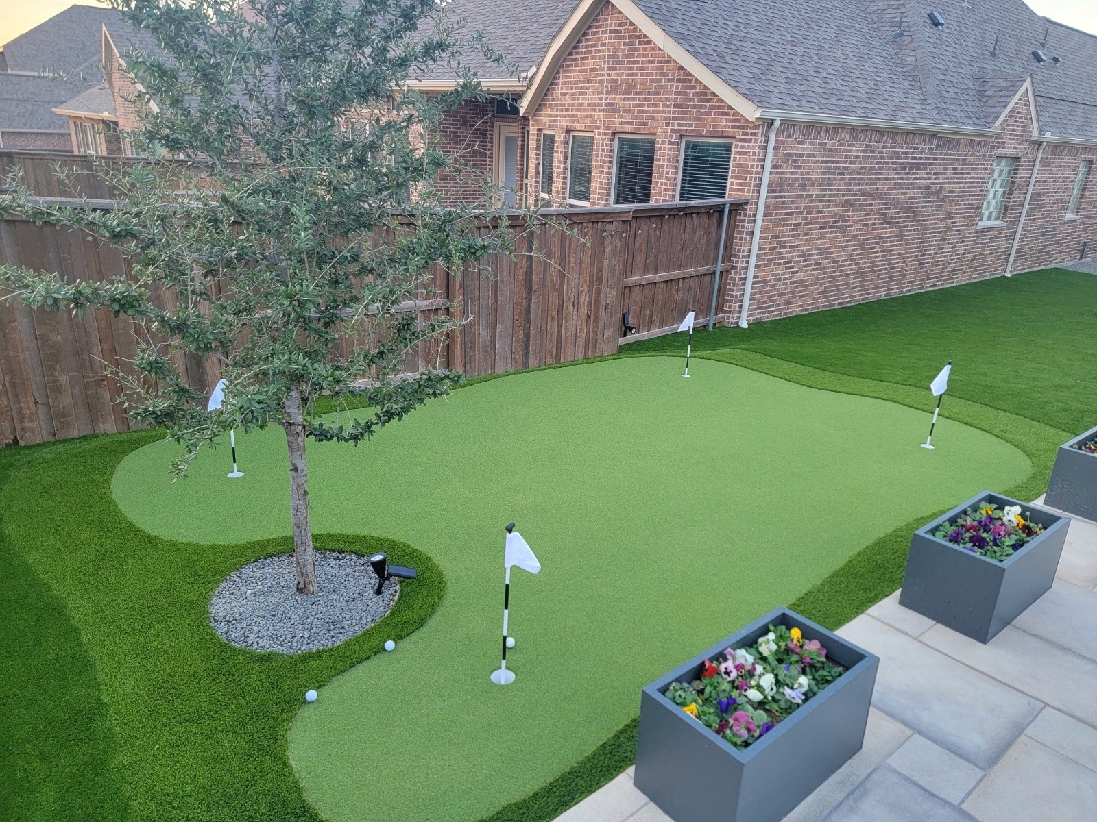 Professionally installed custom backyard putting green made of high-quality artificial turf, featuring five holes, a realistic fringe, and seamlessly integrated around a central tree and paver patio.