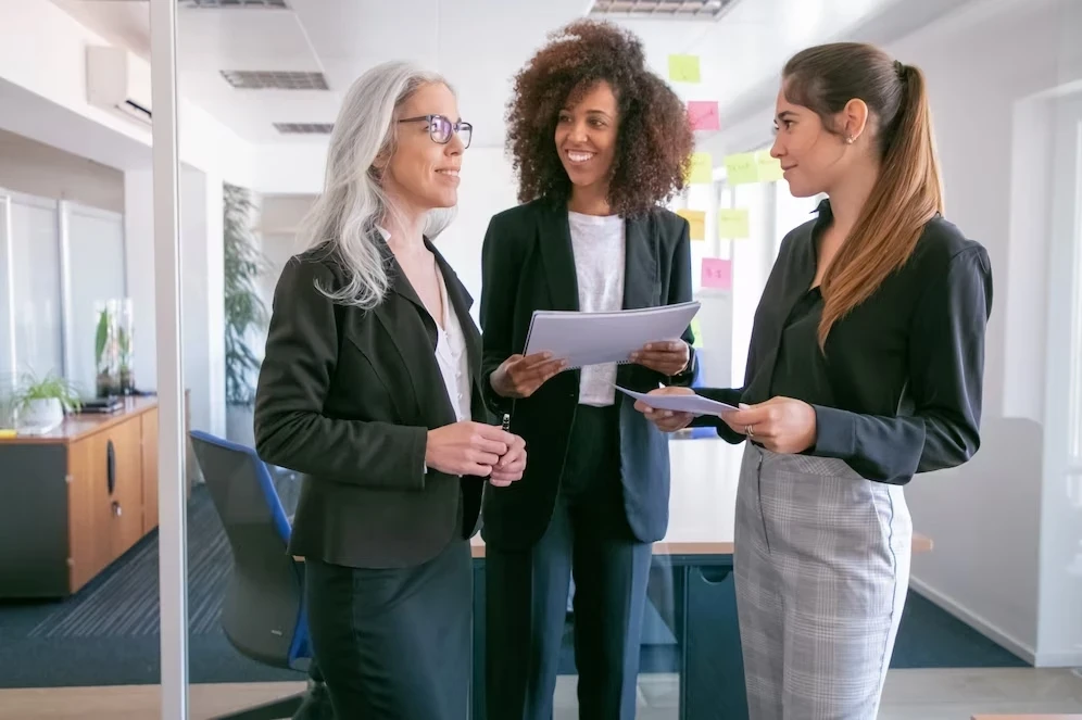 A imagem mostra três mulheres em um ambiente corporativo, todas vestidas formalmente. As três estão interagindo entre si, segurando papéis e sorrindo, o que sugere um momento de colaboração ou discussão amigável. Ao fundo, é possível ver post-its coloridos em uma parede de vidro, indicando um ambiente de trabalho criativo ou um espaço para brainstorming. Essa imagem transmite a ideia de diversidade de gerações e colaboração em um ambiente de trabalho moderno.