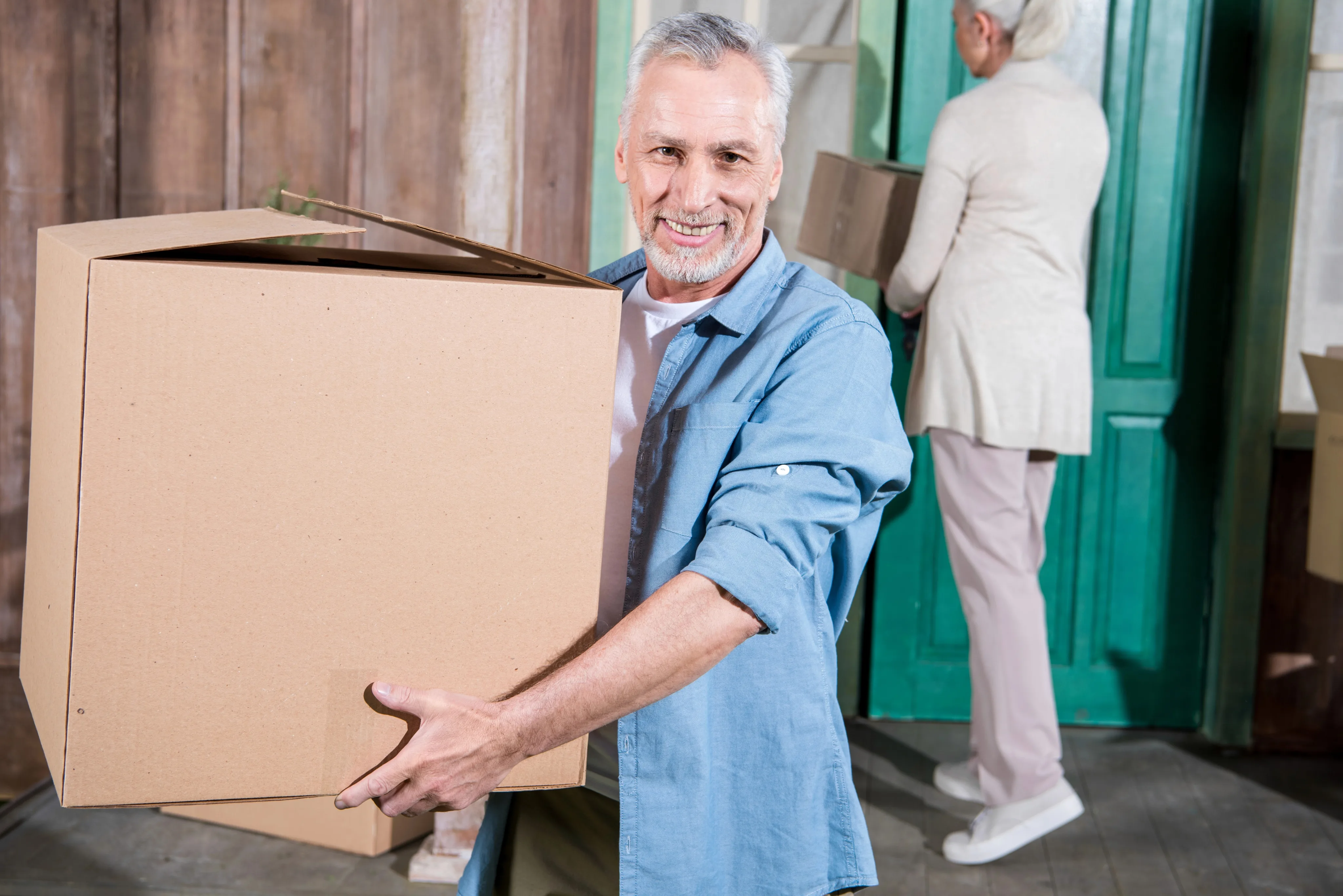 A smiling middle-aged man in a blue shirt carries a large cardboard box while moving, with other people visible in the background near a green door.
