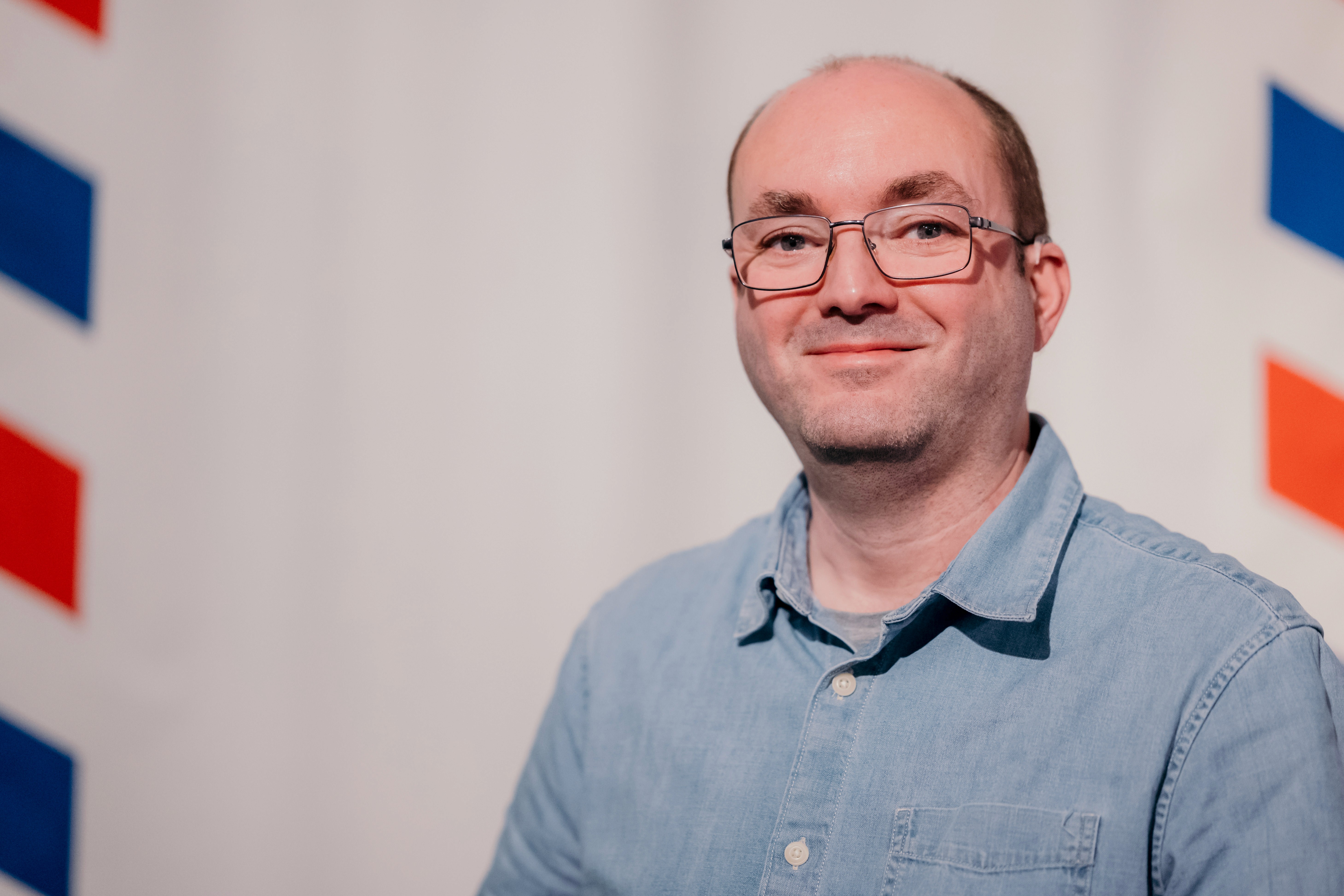 a man with short hair and glasses wearing a light blue shirt smiles at the camera in front of a white background with red and blue patterns to his left and right