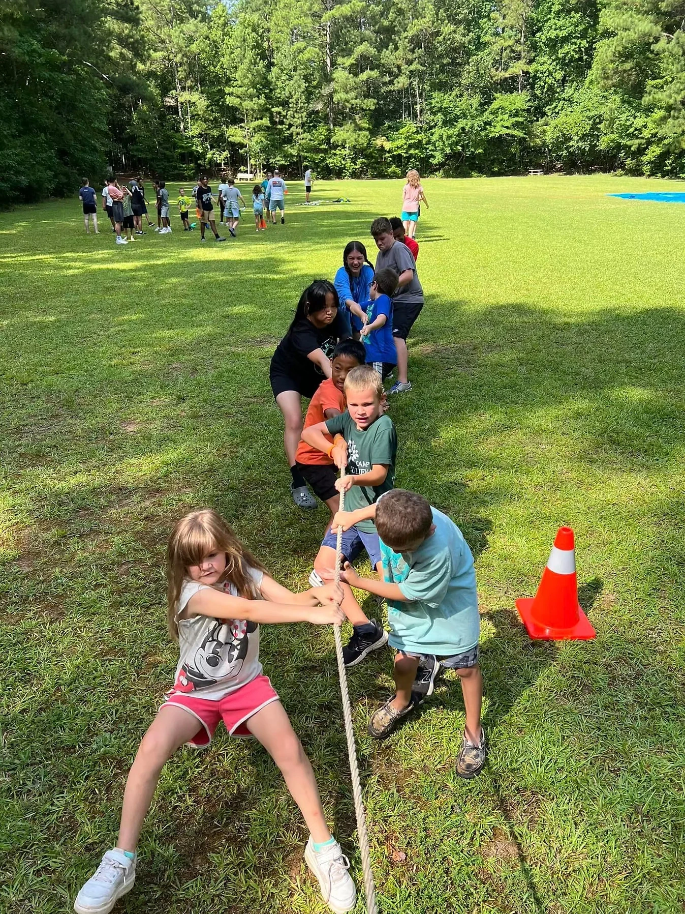 Campers participate in a tug-of-war competition on a grassy field. Two teams pull on a rope, with a bright orange cone nearby.