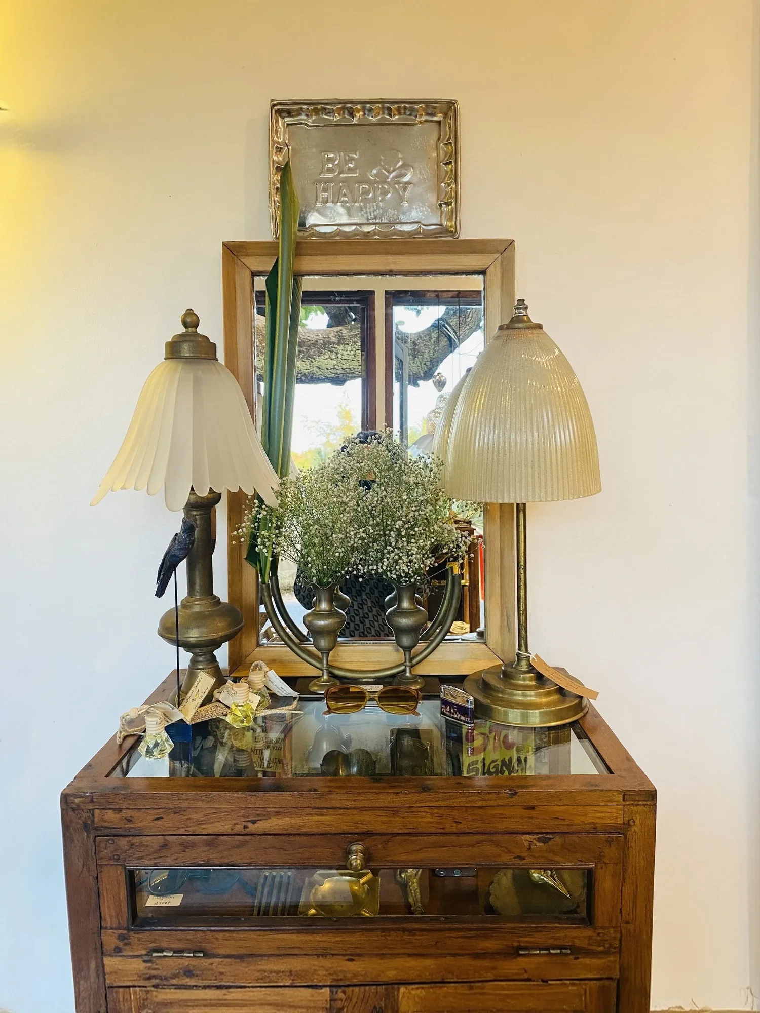 Vintage wooden cabinet with two glass table lamps, an antique mirror and brass vases of baby's breath at Jaali Boutique