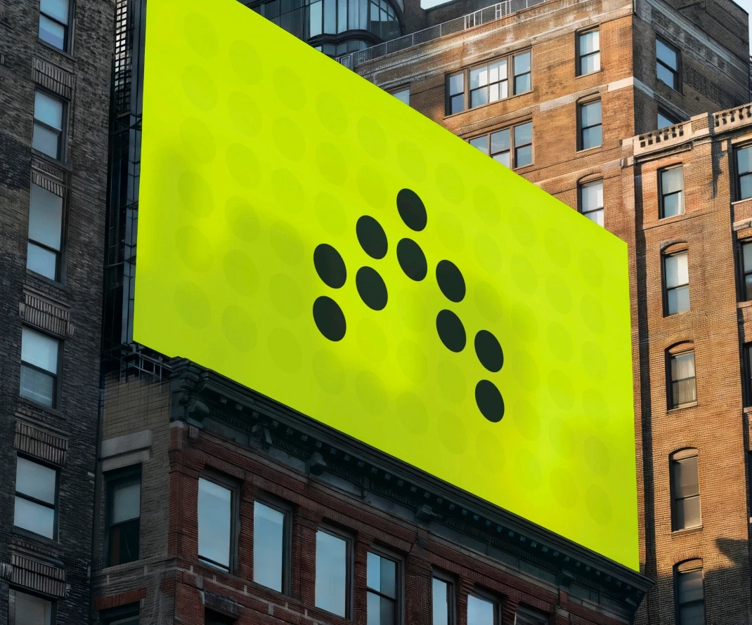 A large, bright green billboard with a minimalist design featuring black dots arranged in a pattern, displayed on the side of a tall urban building. The billboard stands out against the surrounding brick and concrete structures, with windows and architectural details visible.